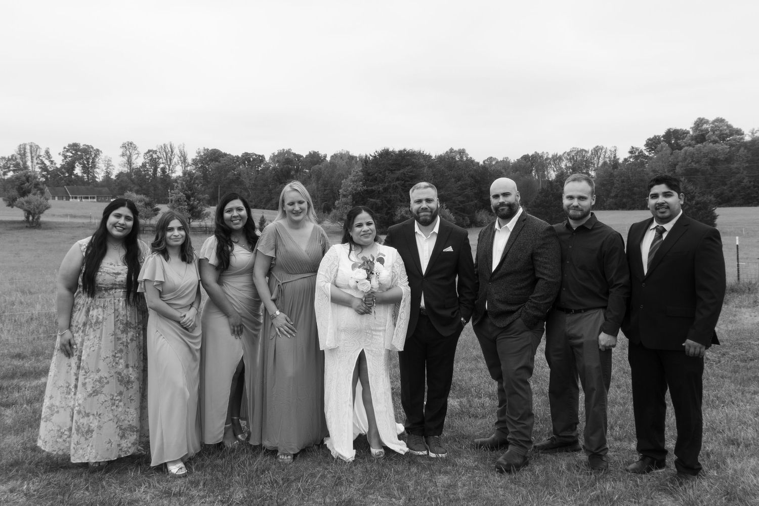 Black and white wedding group photo of nine people posing outdoors in a field, featuring bride and groom in center.