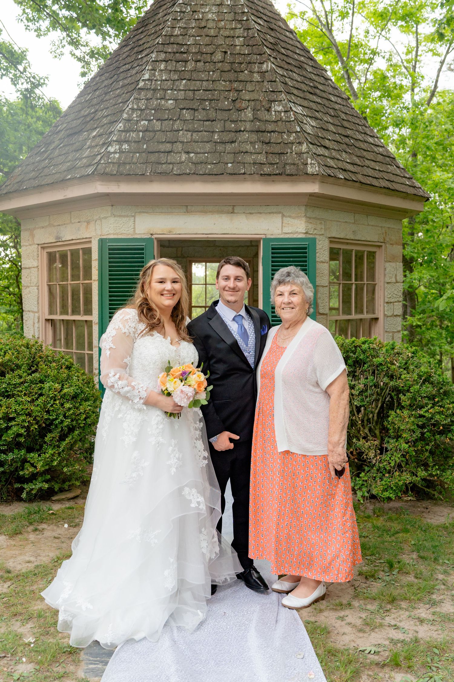 Bride in white lace gown holding bouquet poses with groom in suit and older woman in peach dress before stone gazebo.