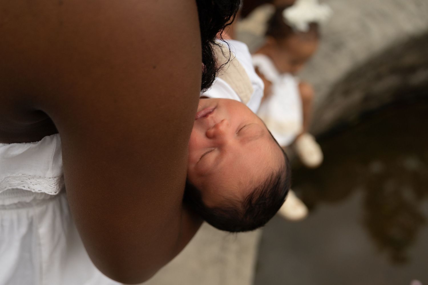 A mother tenderly holds her sleeping newborn baby dressed in white outdoors near water.