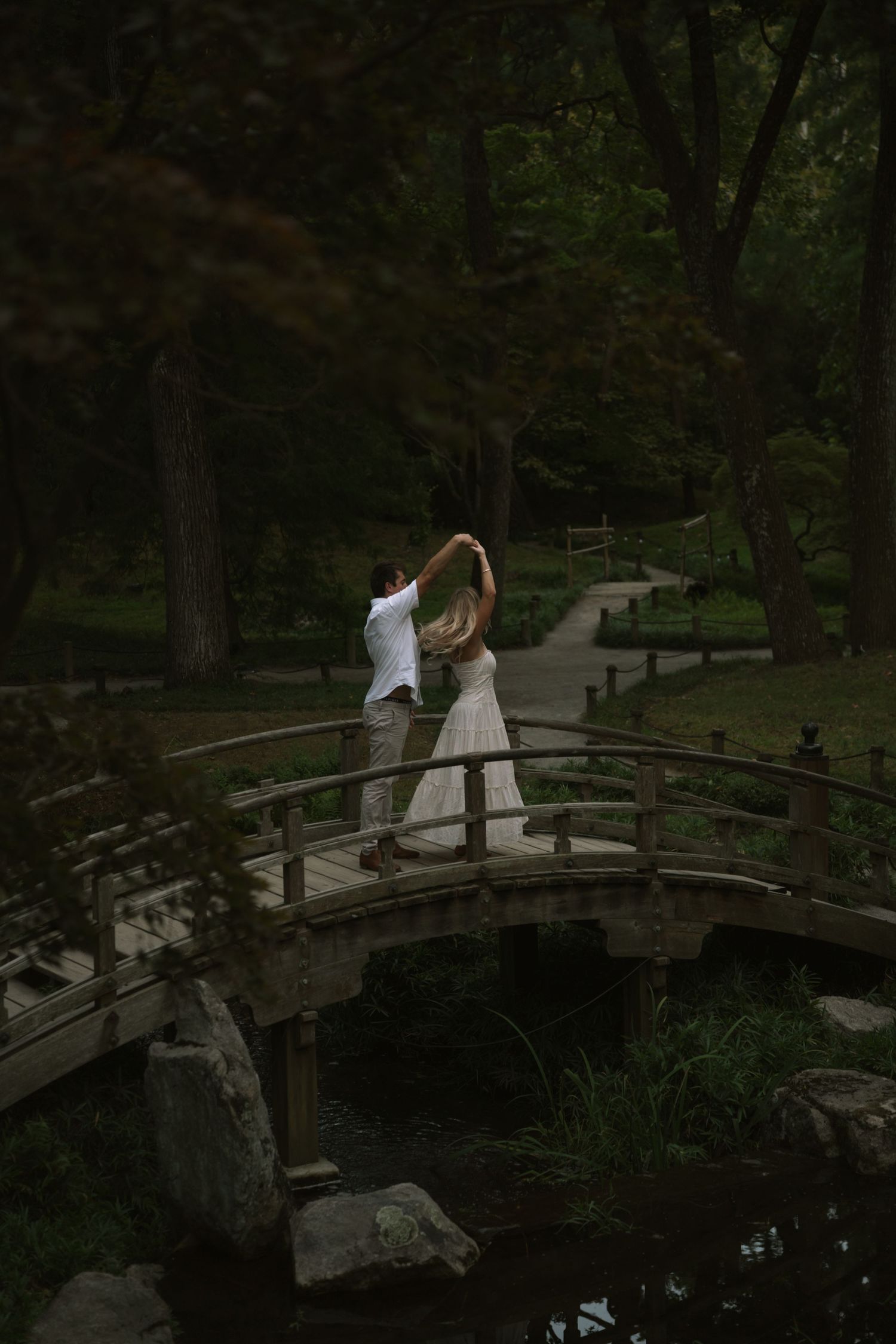 Couple dancing on wooden bridge in lush green garden, bride in white dress, groom in white shirt, romantic outdoor setting.