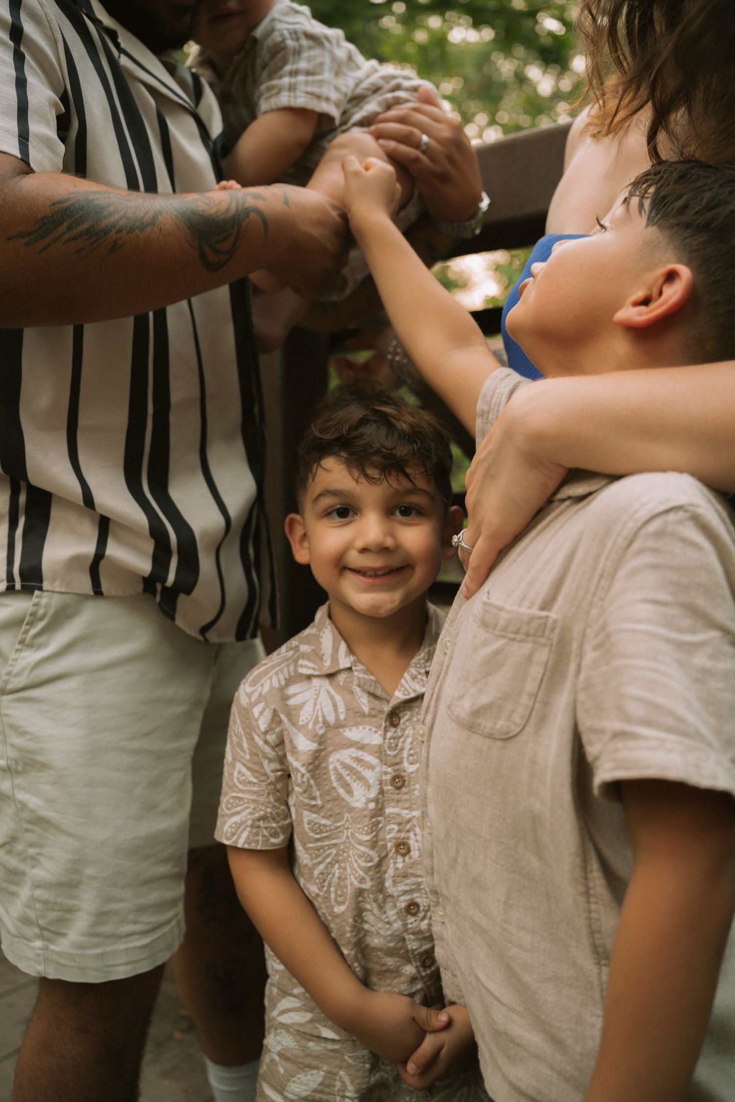 Young boy smiling up at family members outdoors, surrounded by adults and a sibling in a casual summer setting. Row and Co Photography