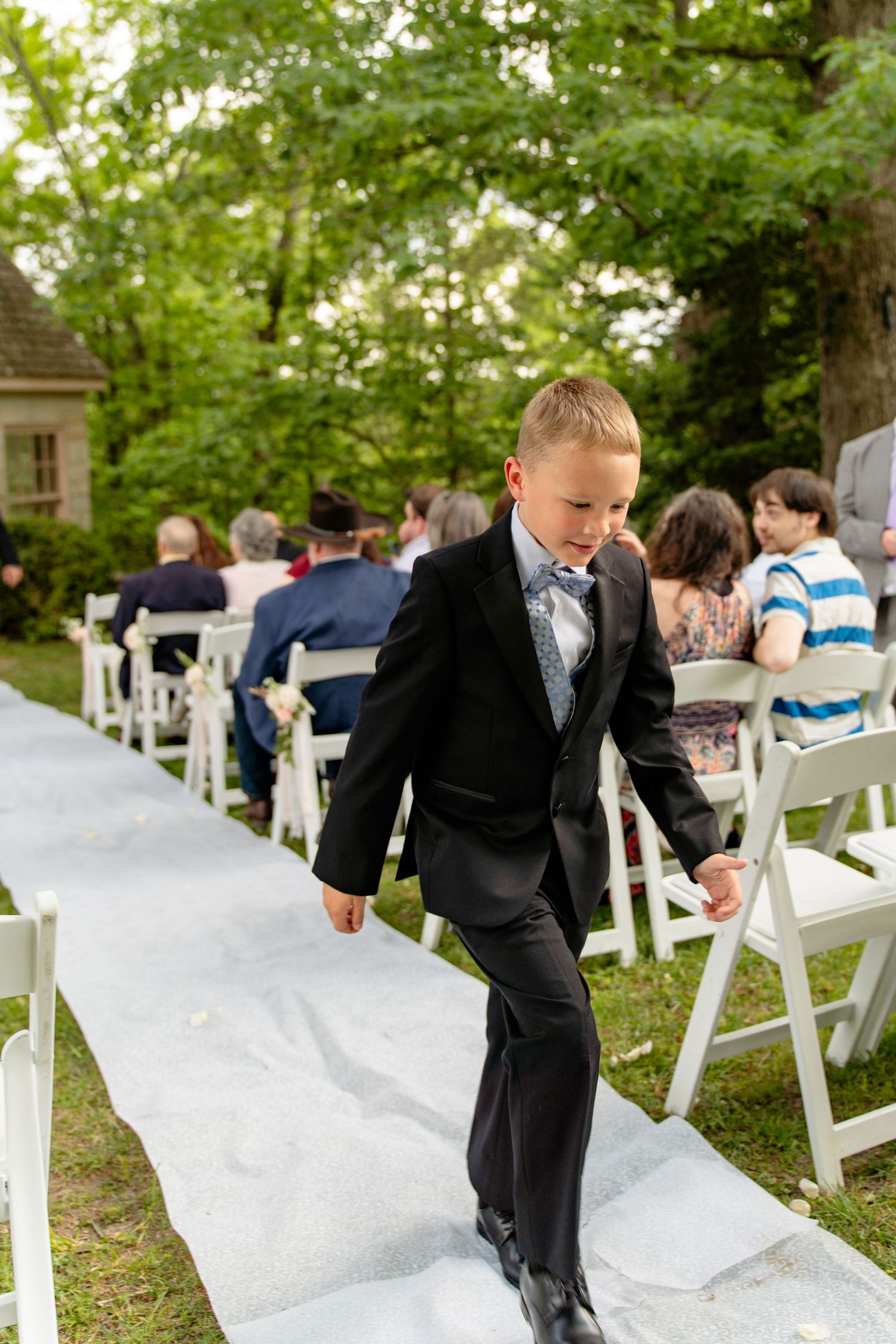 Young boy in black suit walks down white aisle at outdoor wedding ceremony with guests seated on white chairs.