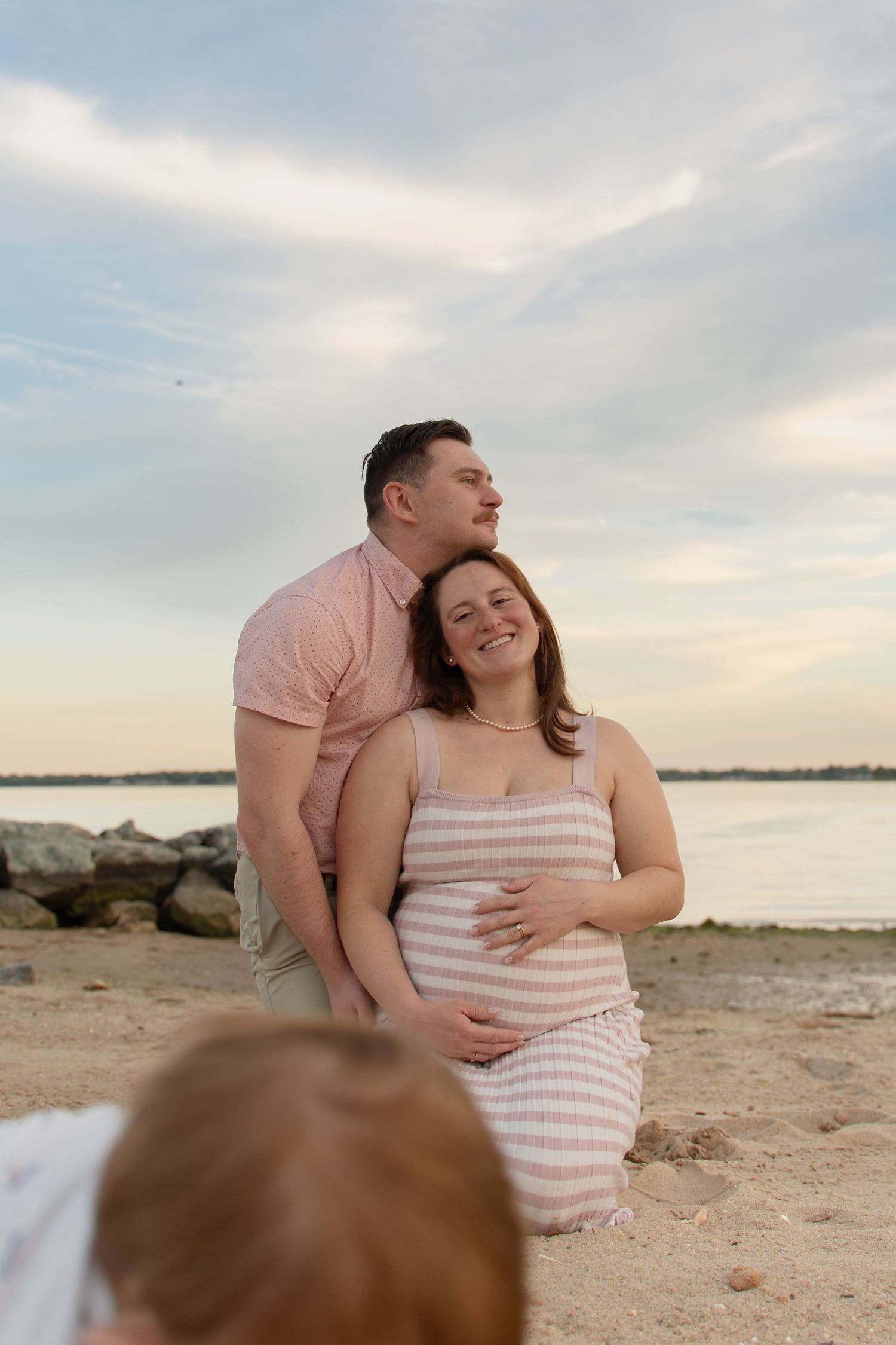 A couple poses on a sandy beach at sunset, the pregnant woman smiling as her partner kisses her head.