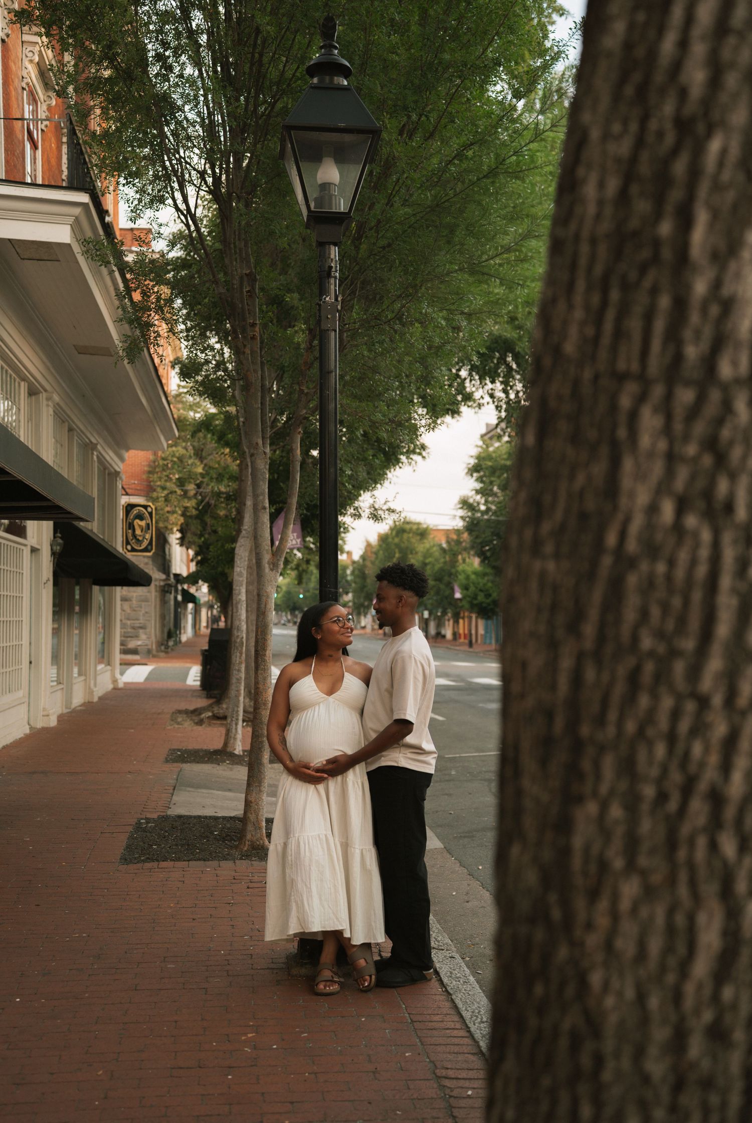 Pregnant couple in white outfits share a tender moment on a tree-lined urban sidewalk near a vintage street lamp.