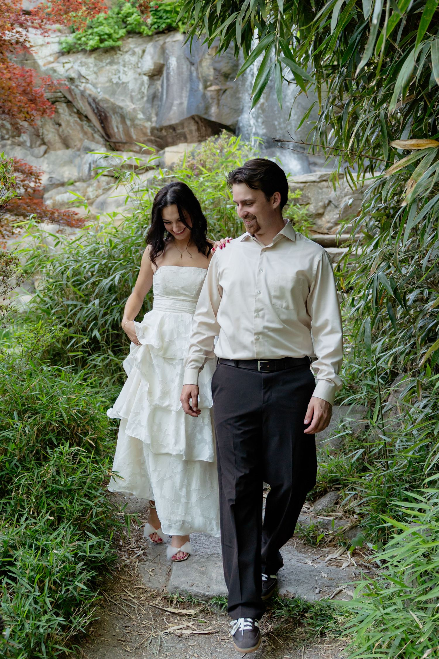 Couple in wedding attire walking hand in hand along a garden path surrounded by lush greenery and a waterfall. Taken by Row And Co