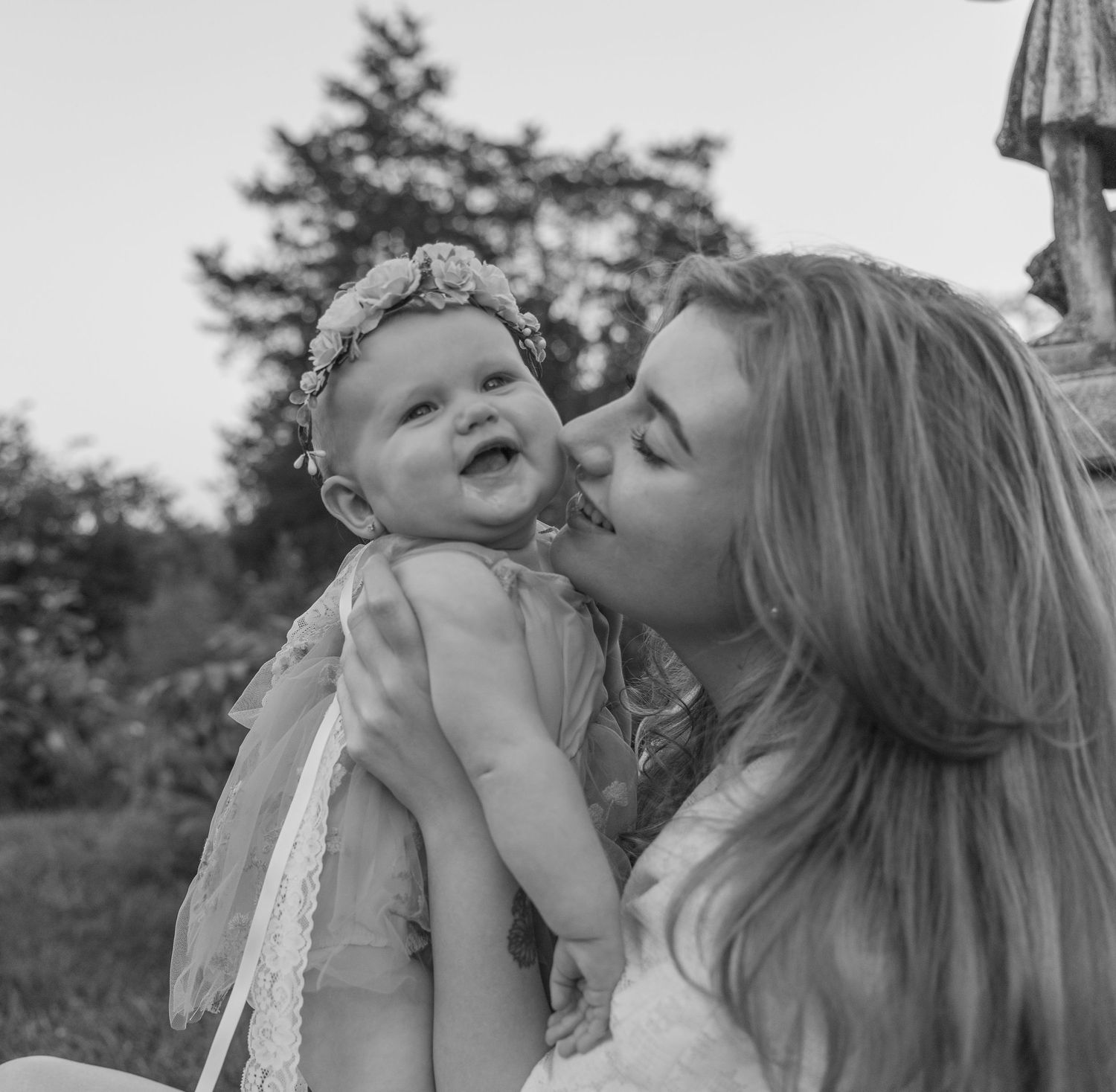 Black and white photo of a young woman kissing a smiling baby girl wearing a floral headband outdoors.