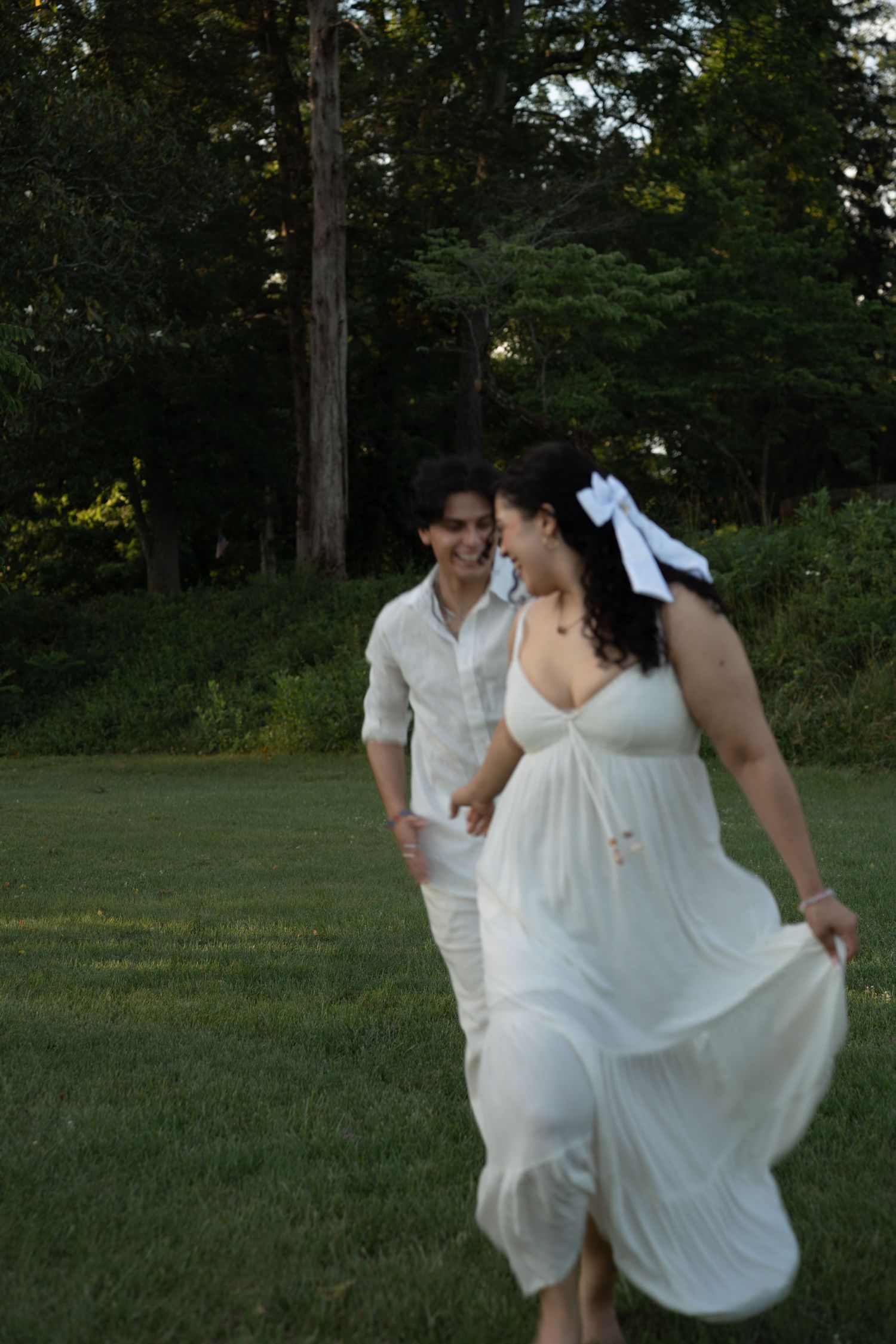 A smiling couple in white attire runs through a lush green lawn surrounded by trees at dusk.