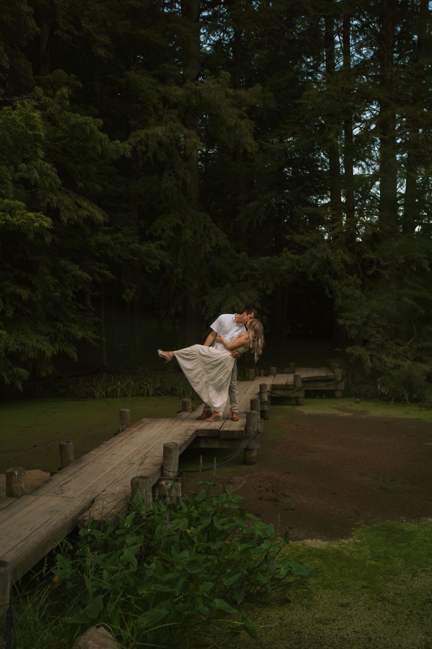 Couple sharing a romantic dip kiss on a wooden bridge surrounded by lush green trees in a serene garden setting.