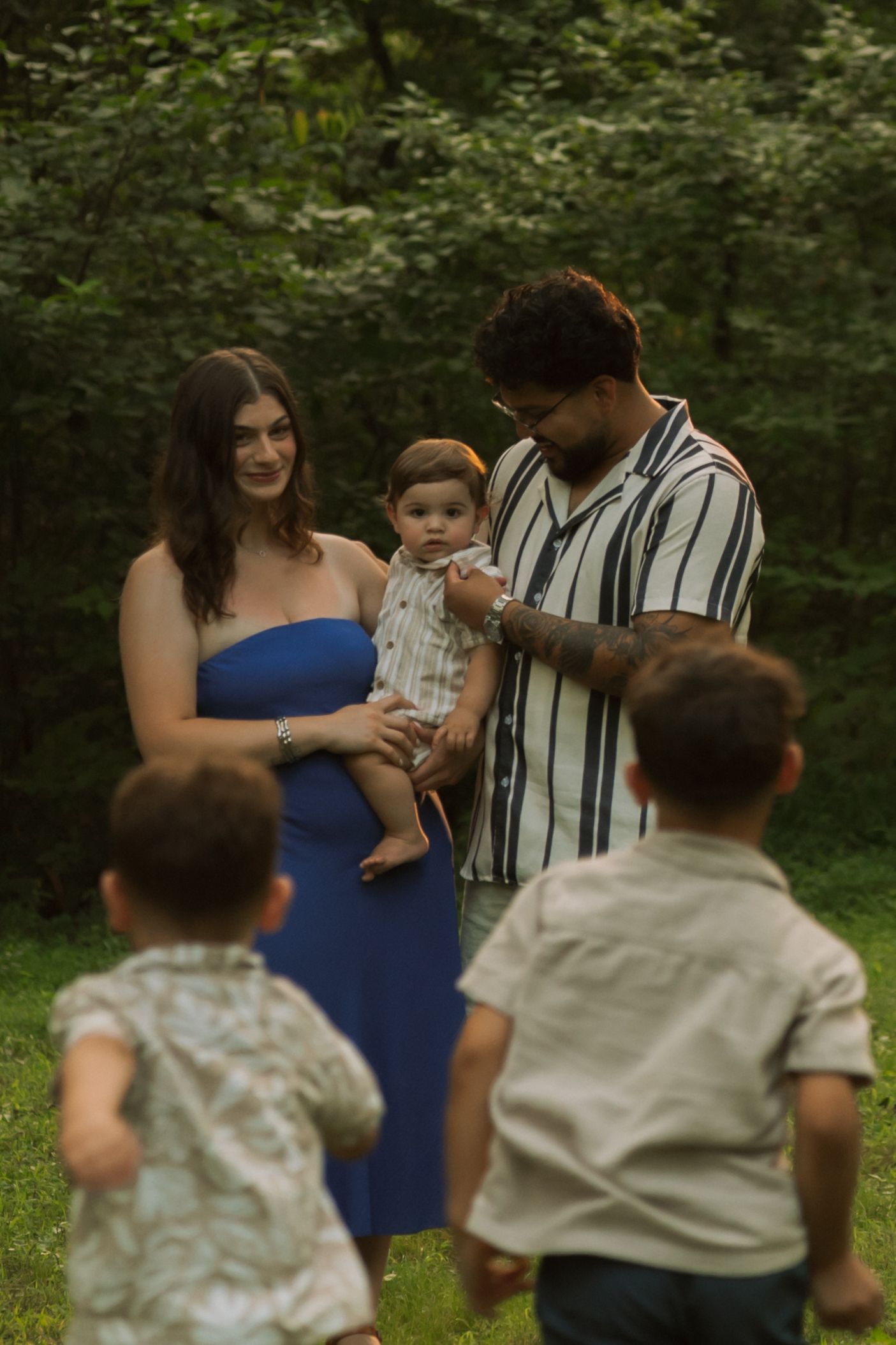 Family of four outdoors, parents holding baby while two young children run toward them in a lush green setting.