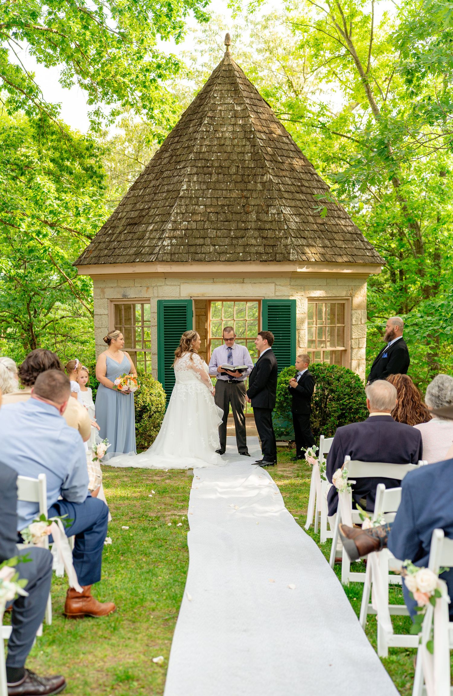 Outdoor wedding ceremony at a charming stone gazebo, bride and groom exchange vows before seated guests on a white aisle.