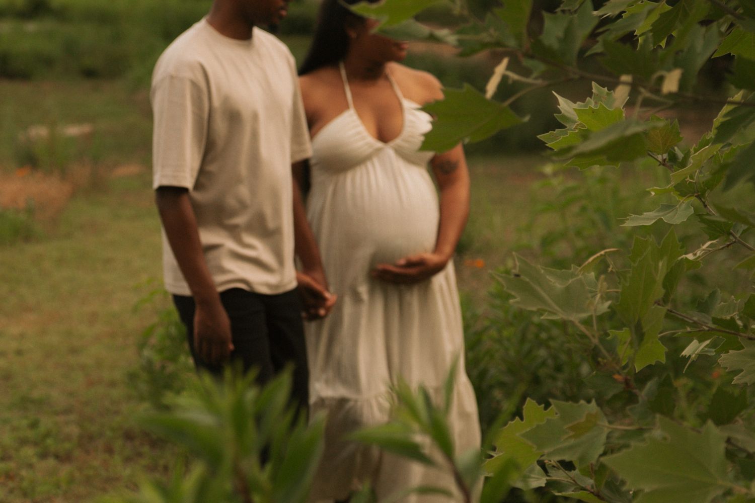 Pregnant Black couple in white outfits standing together outdoors surrounded by lush green foliage during maternity photoshoot.
