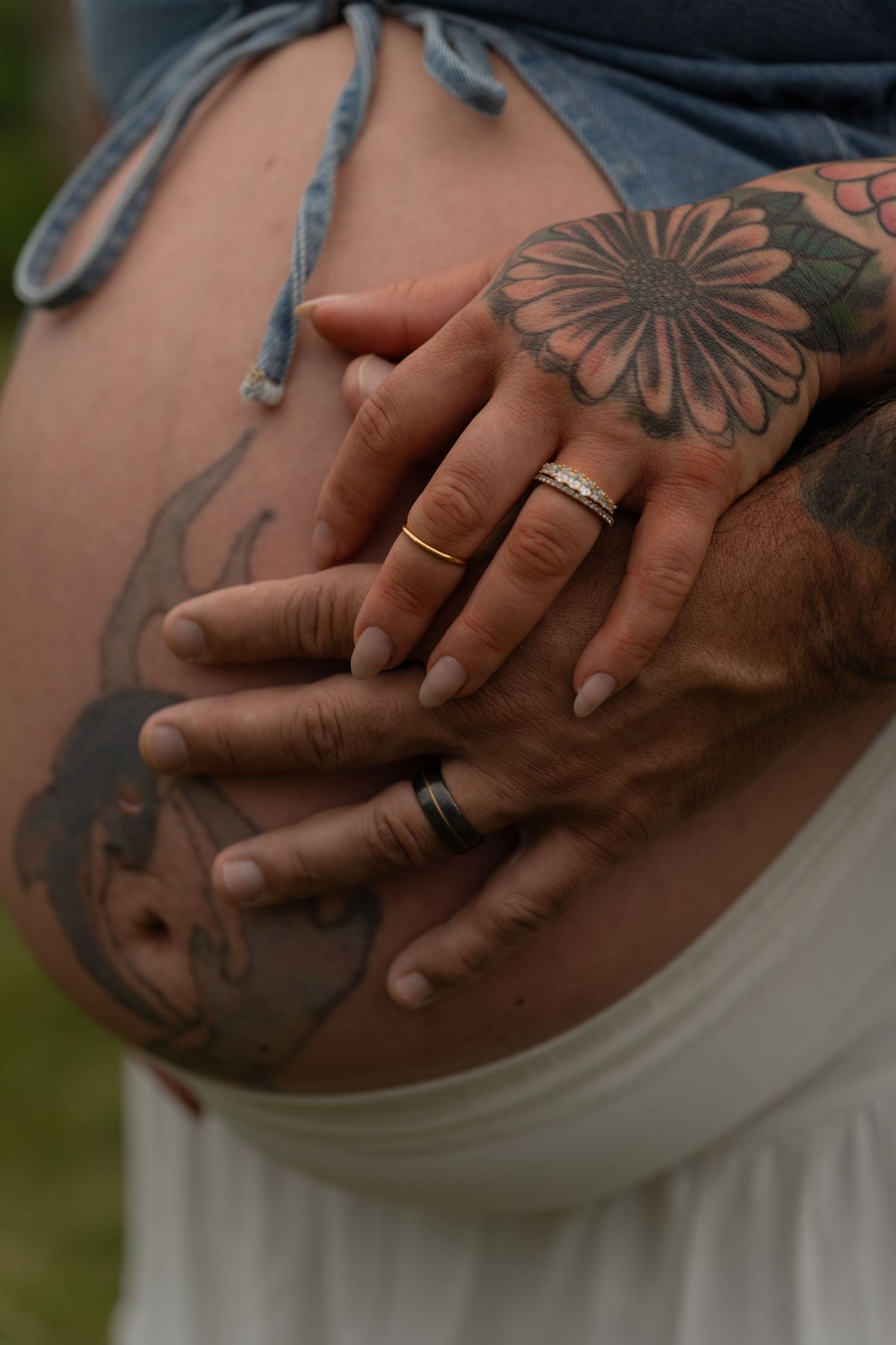 Couple placing tattooed hands on pregnant belly, wearing wedding rings, with floral tattoo visible.