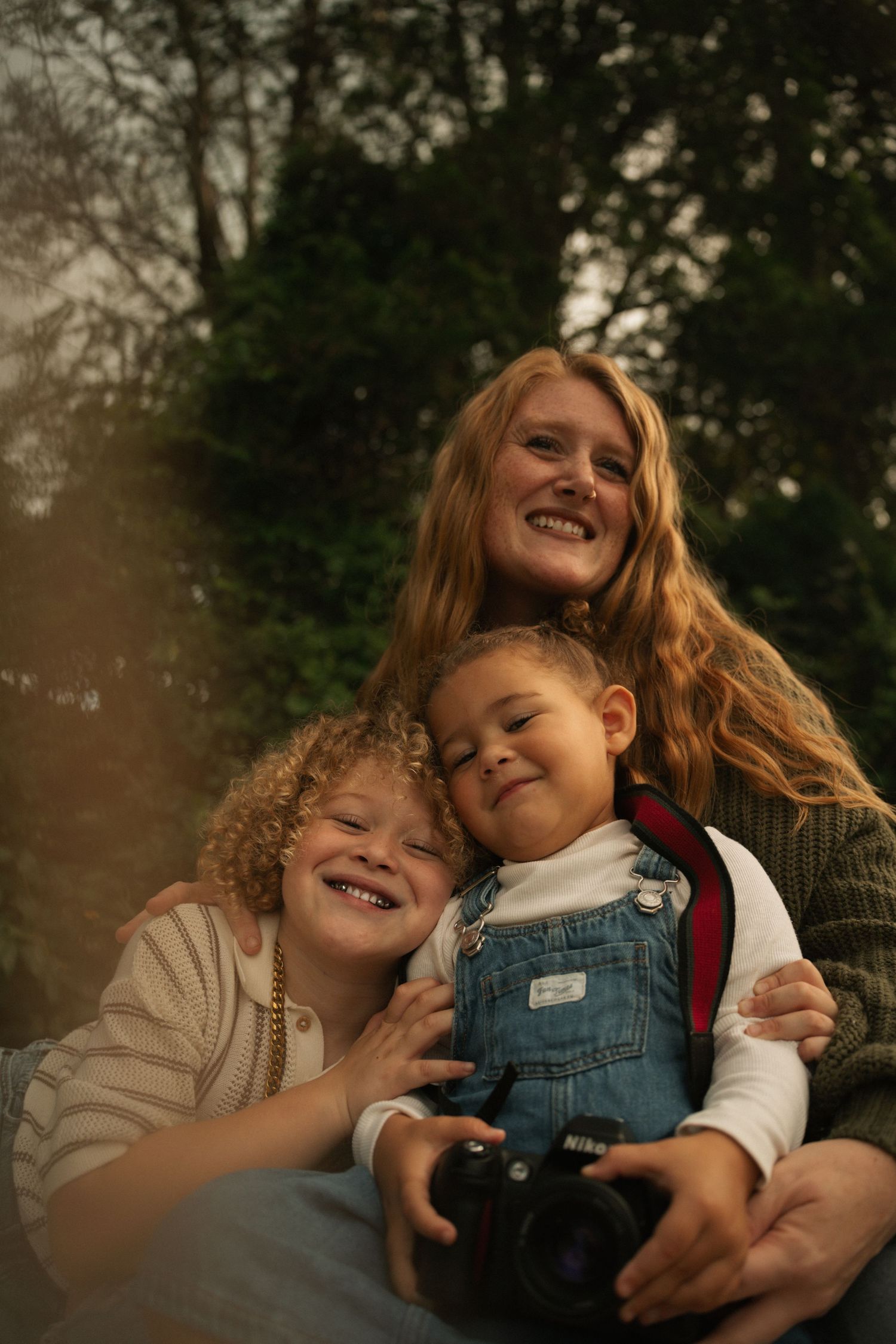 A smiling woman with red hair hugs two young children outdoors, one in denim overalls holding a camera.