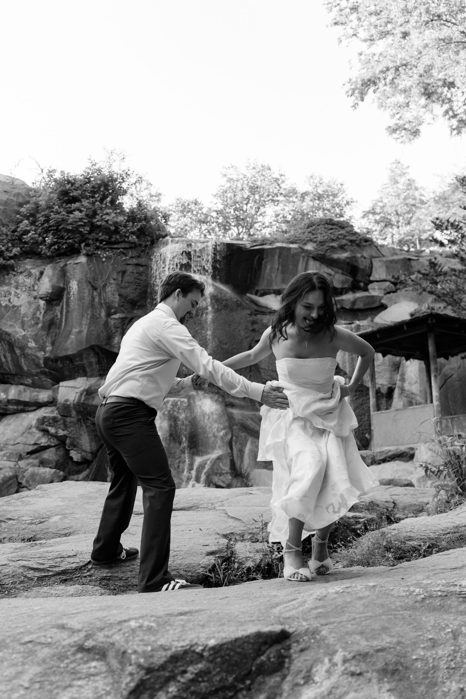 Black and white photo of groom helping bride navigate rocky terrain near waterfall at wedding. Taken by Row And Co Photography in FXBG VA