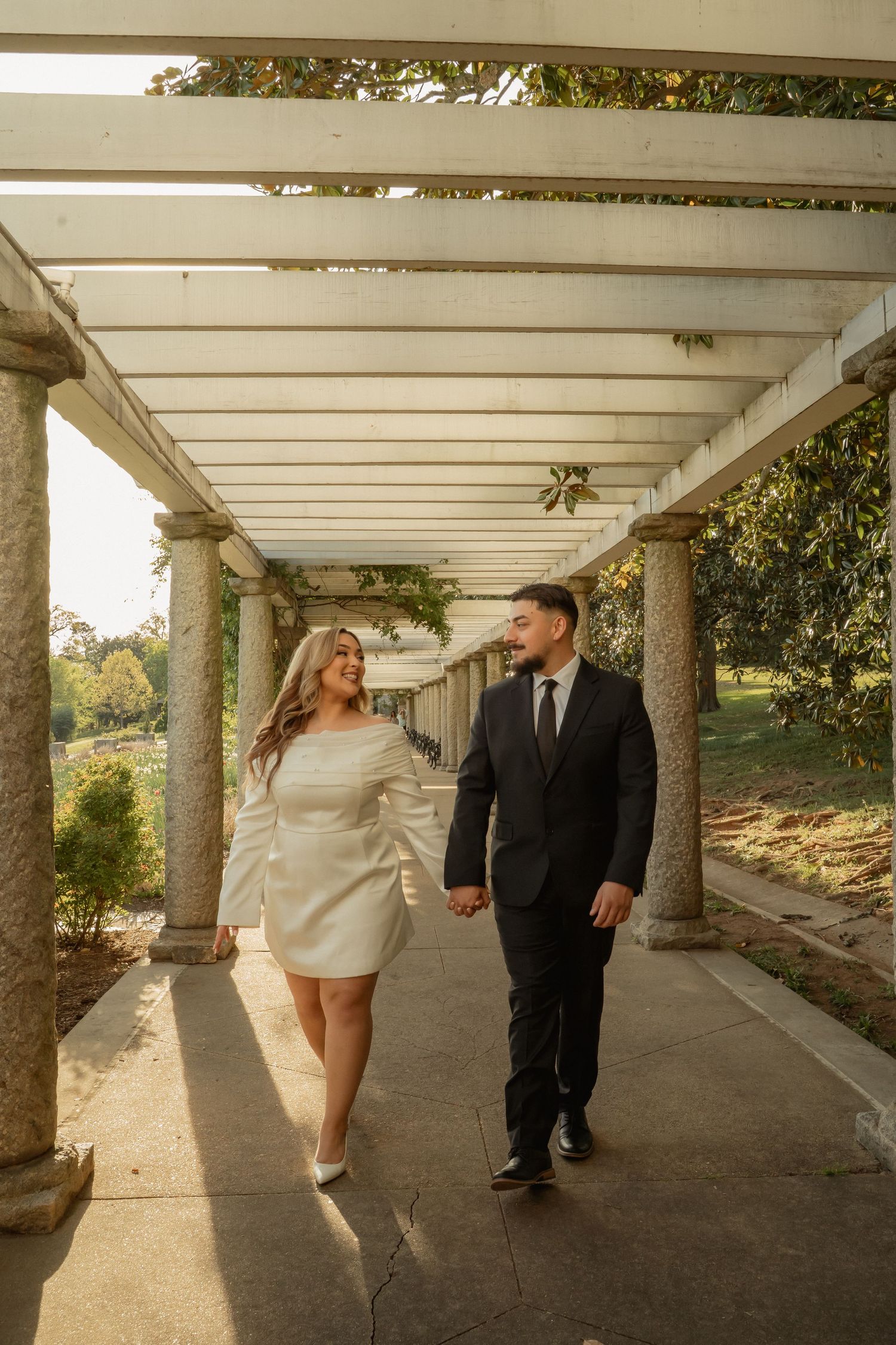 Couple in formal attire holding hands while walking under a white pergola surrounded by lush greenery.