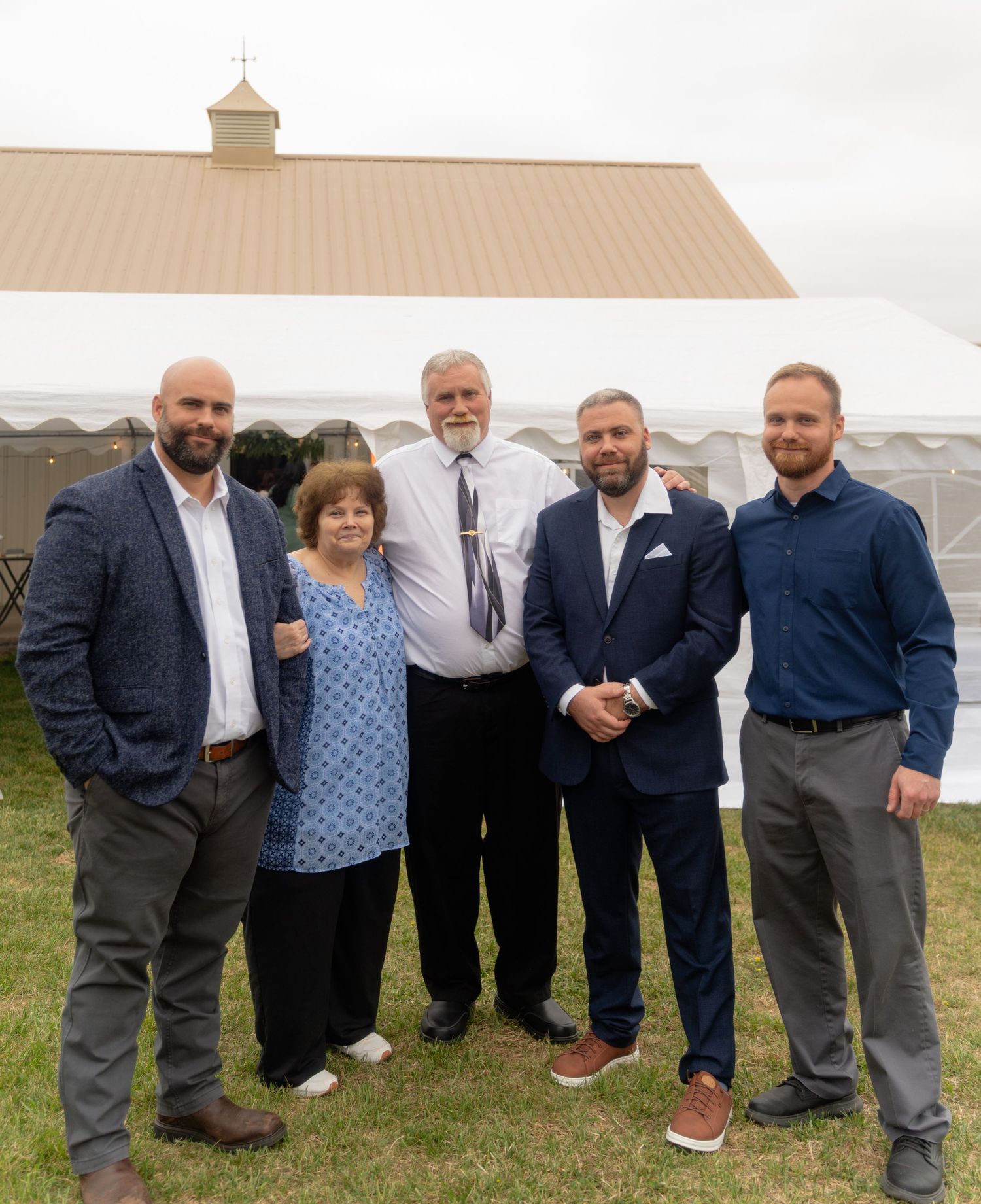 Group of five adults posing outdoors near a white tent and barn at a formal gathering or celebration.