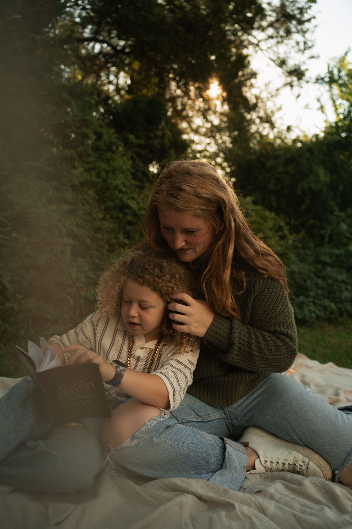 A young woman with red hair reads a book with a curly-haired toddler on a blanket outdoors at golden hour.