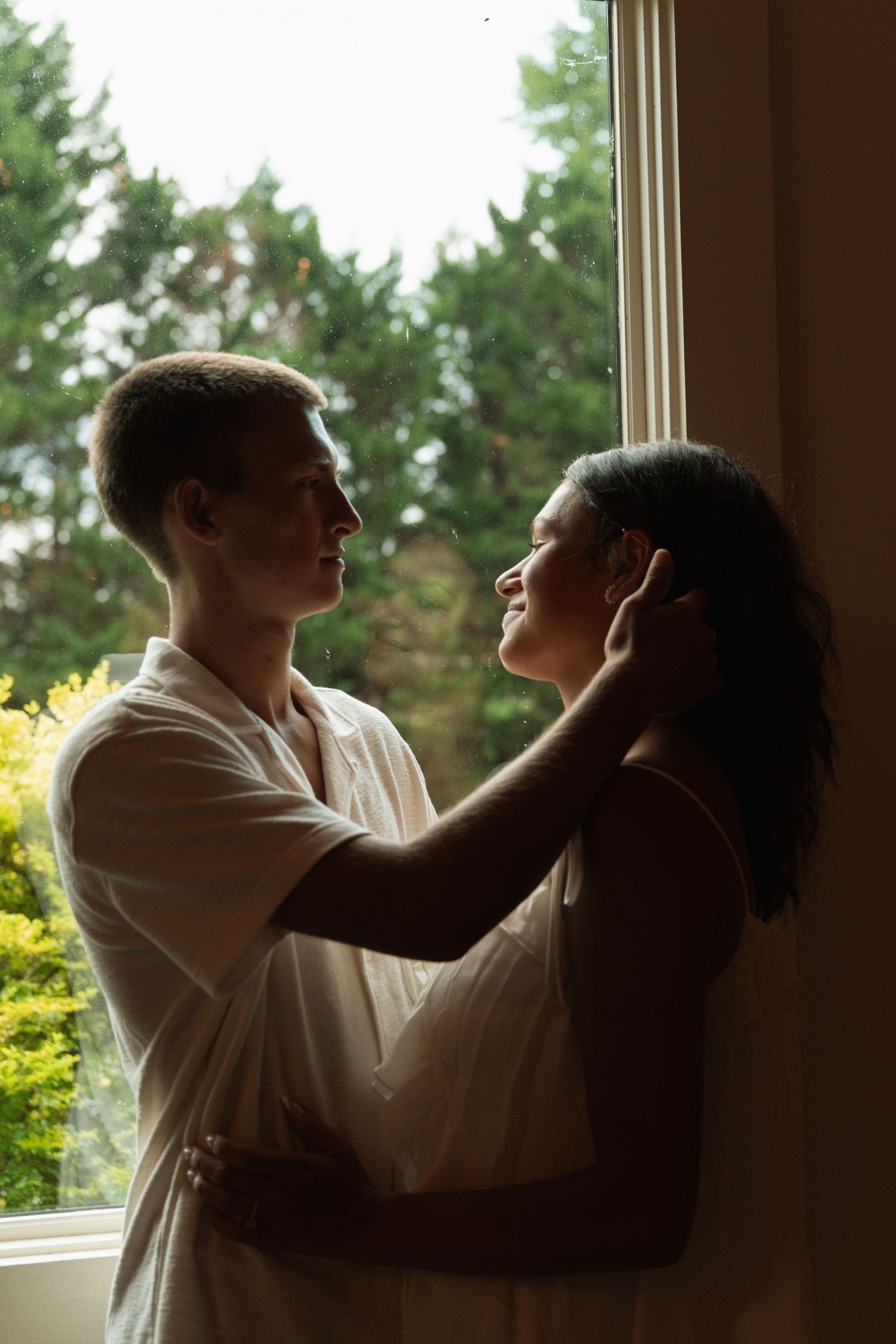 Romantic couple embracing by a sunlit window with lush green trees in the background.
