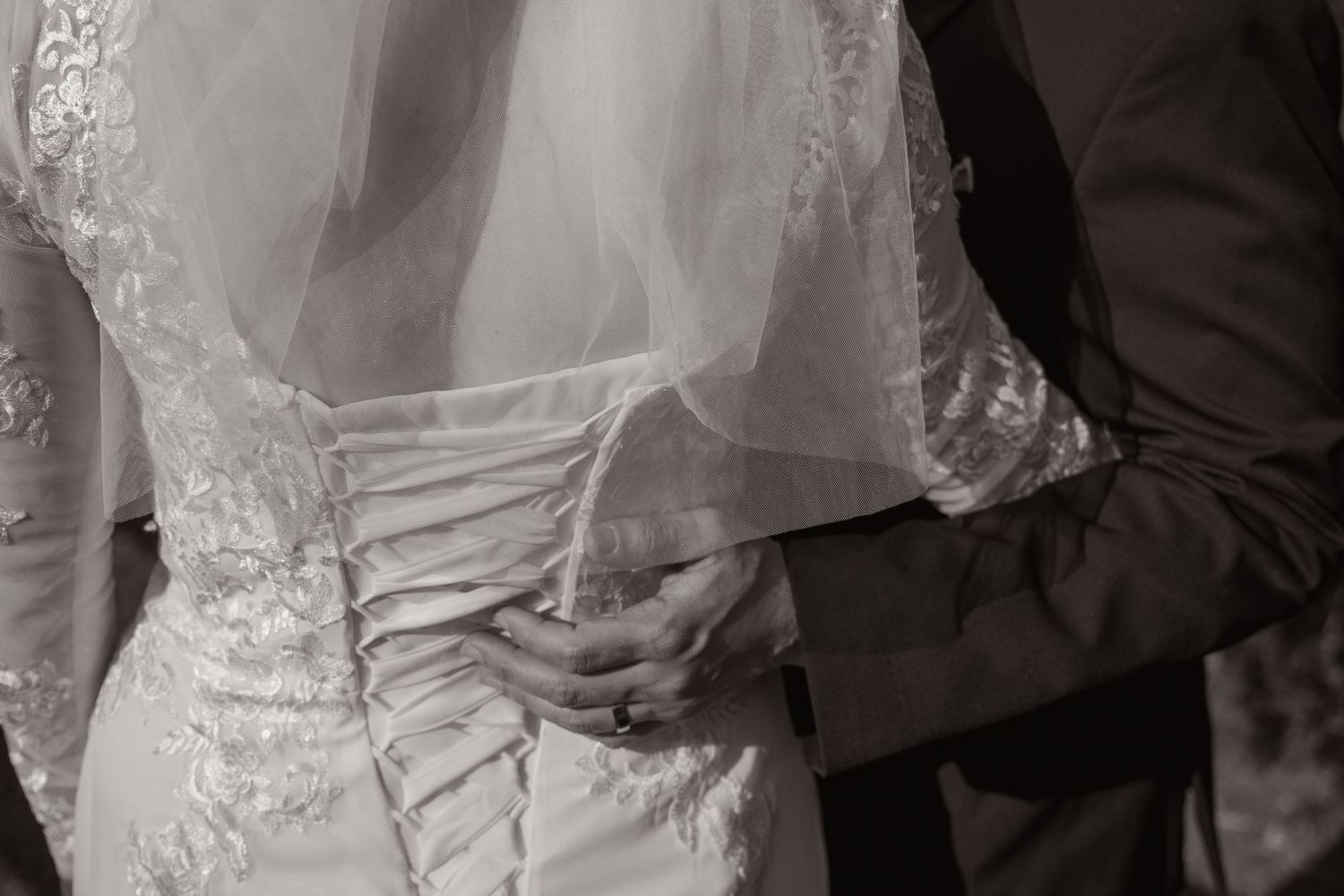 Black and white photo of bride and groom embracing, showcasing lace wedding dress back and veil.