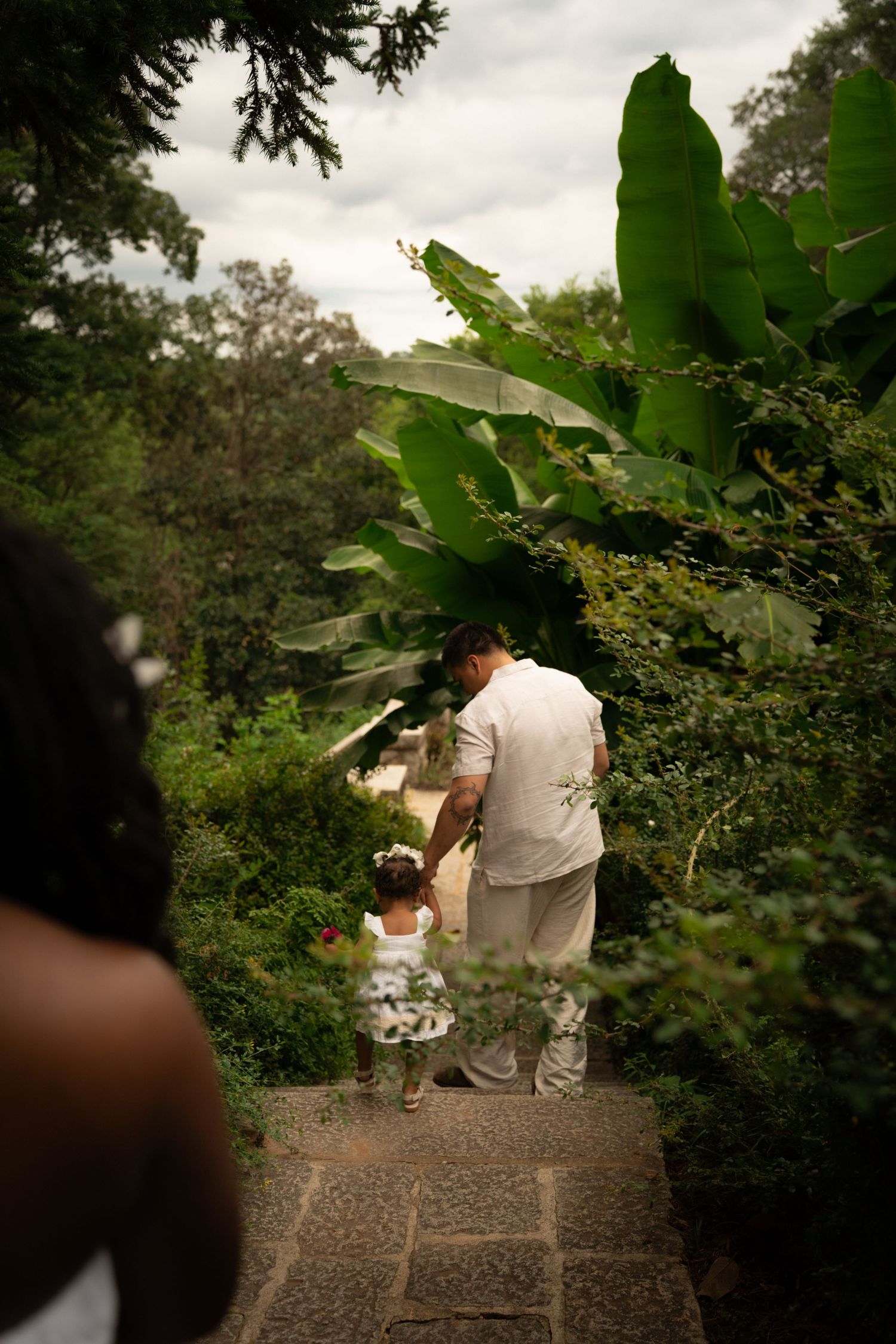Adult and child walking hand in hand along a stone path surrounded by lush tropical garden greenery.