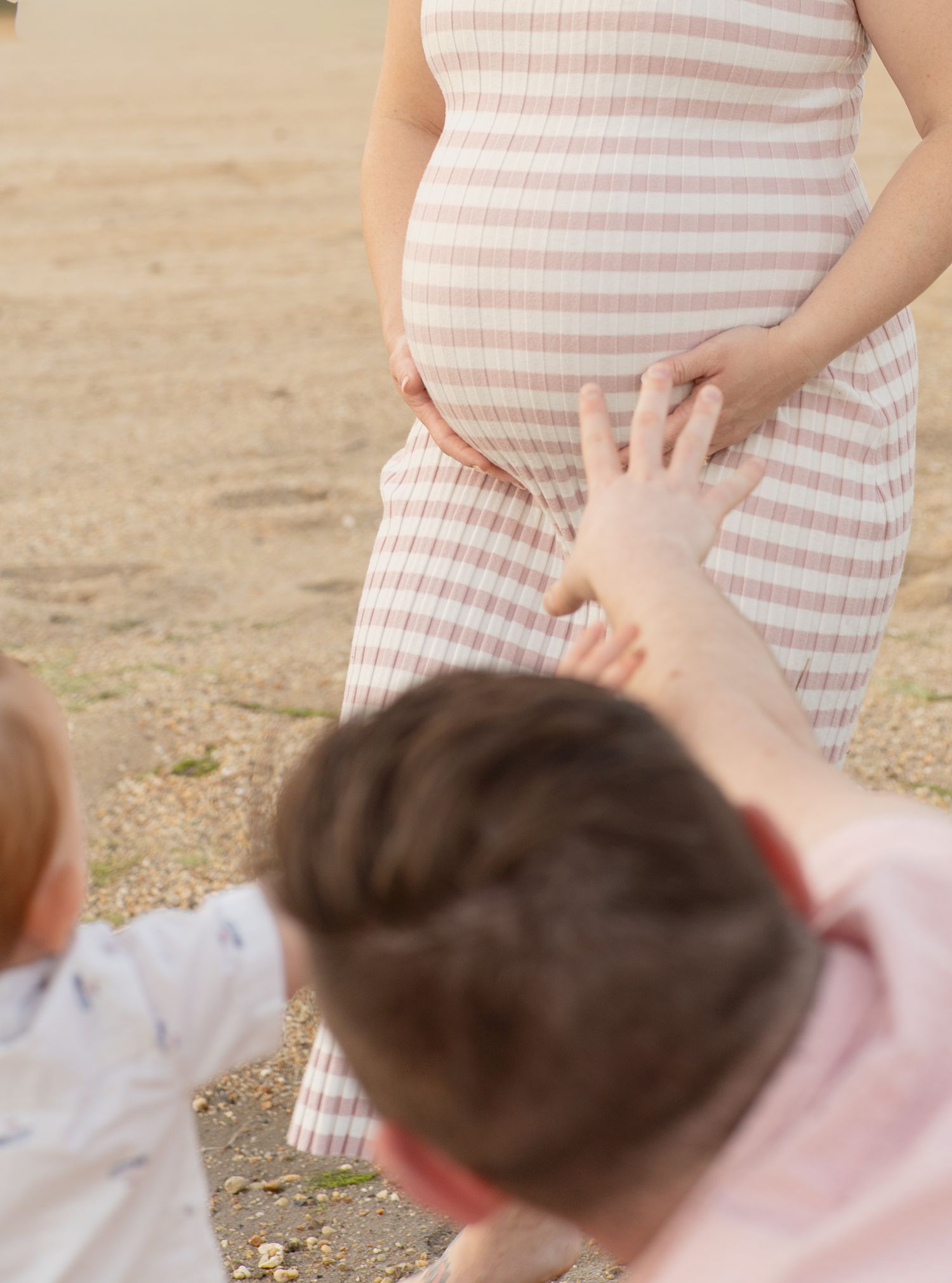 Pregnant woman in striped dress with children touching her baby bump outdoors.