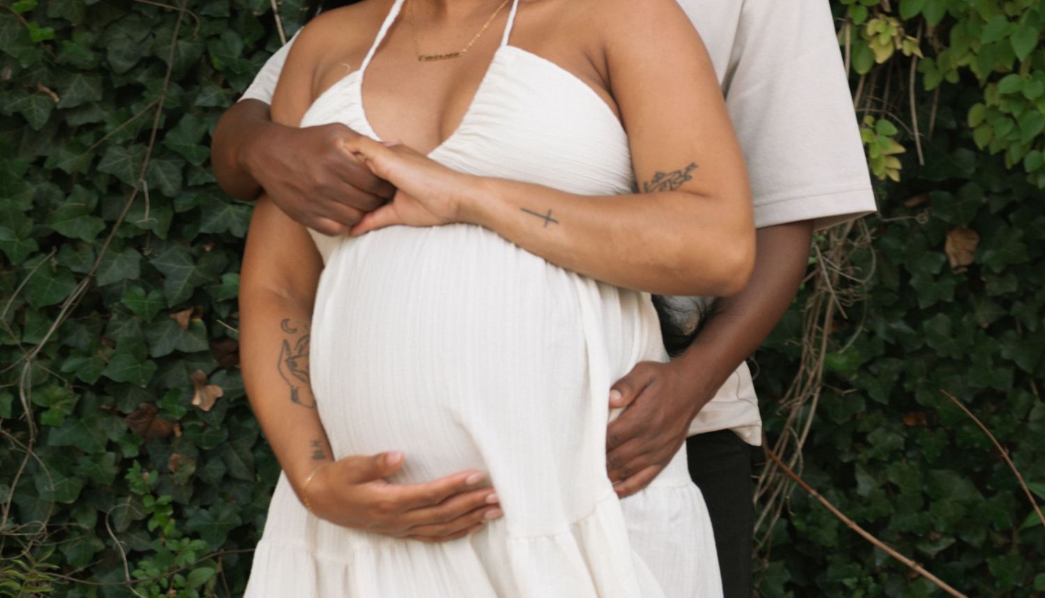 Close-up of a pregnant woman in white dress with partner's tattooed arms cradling her baby bump outdoors.