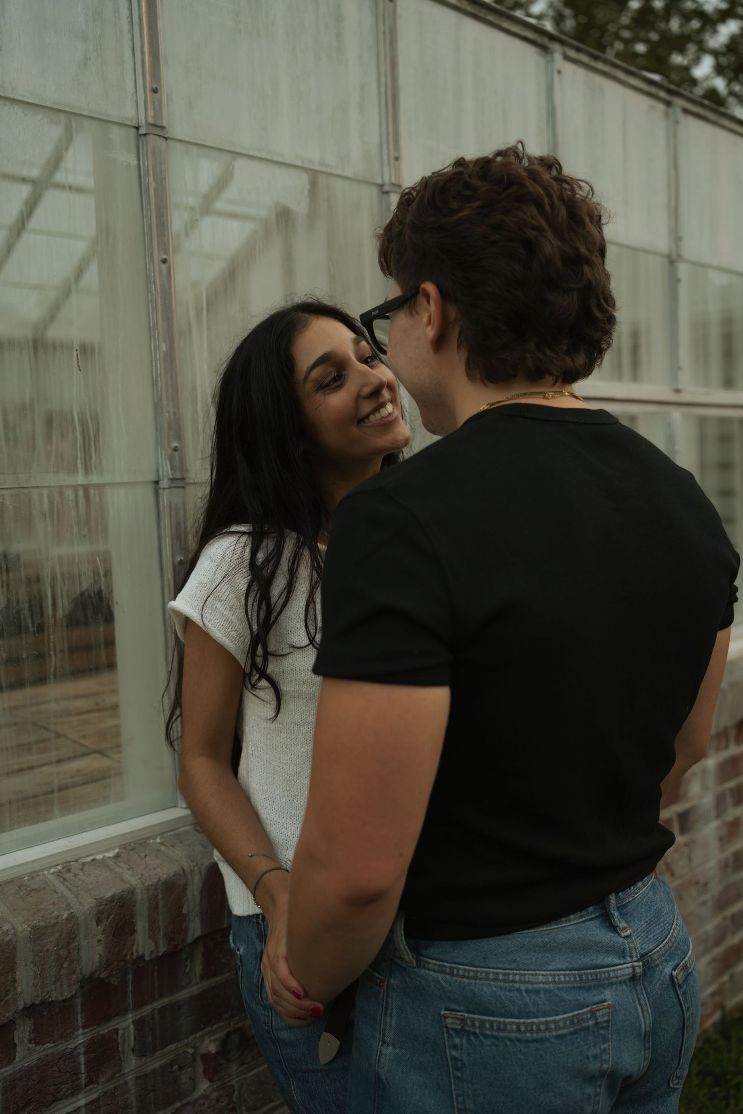 A smiling young woman in a white tee holds hands with a man in a black shirt near a greenhouse wall.