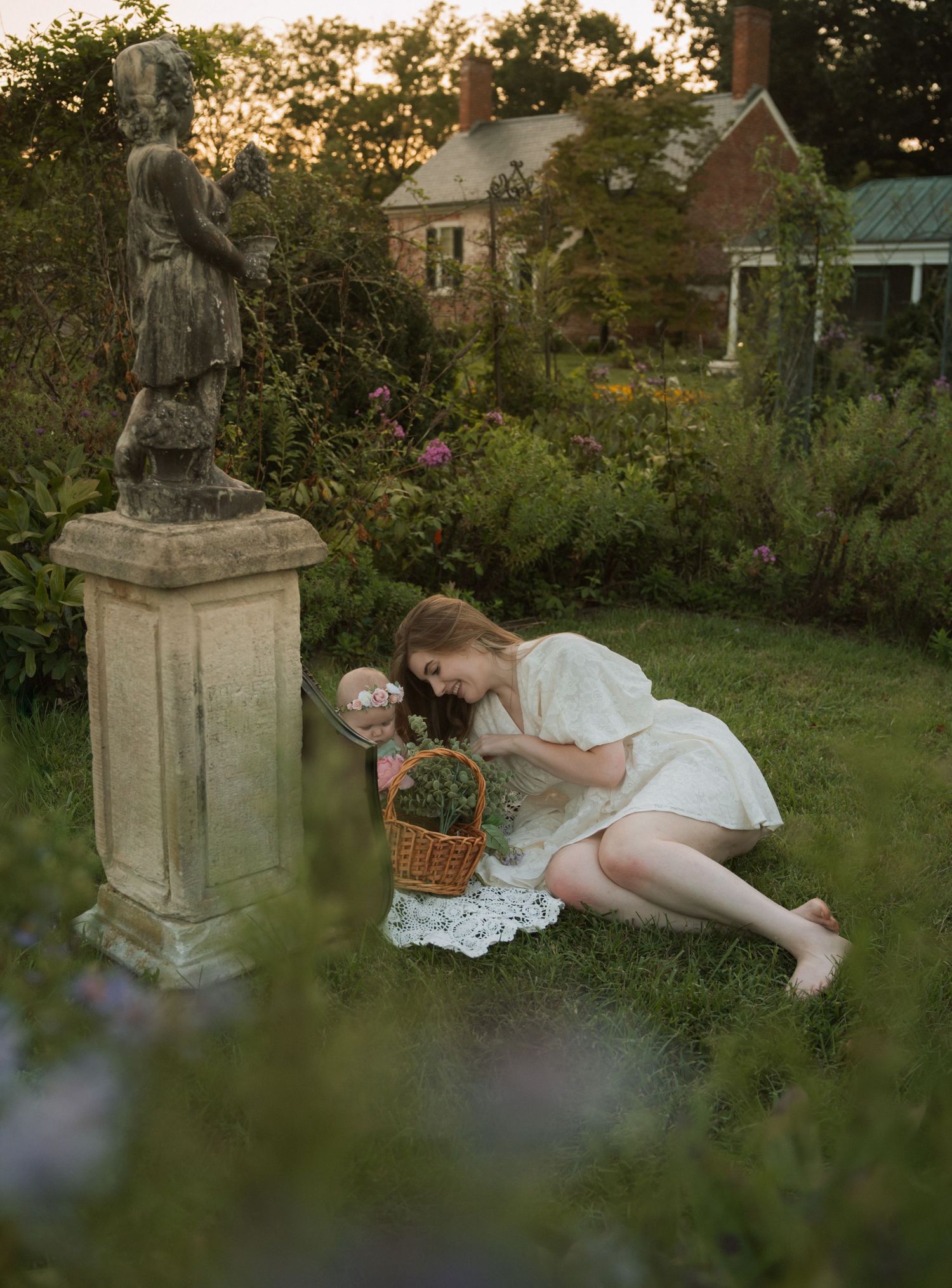 Woman in white dress kneeling in lush garden beside stone statue, holding wicker basket at golden hour.