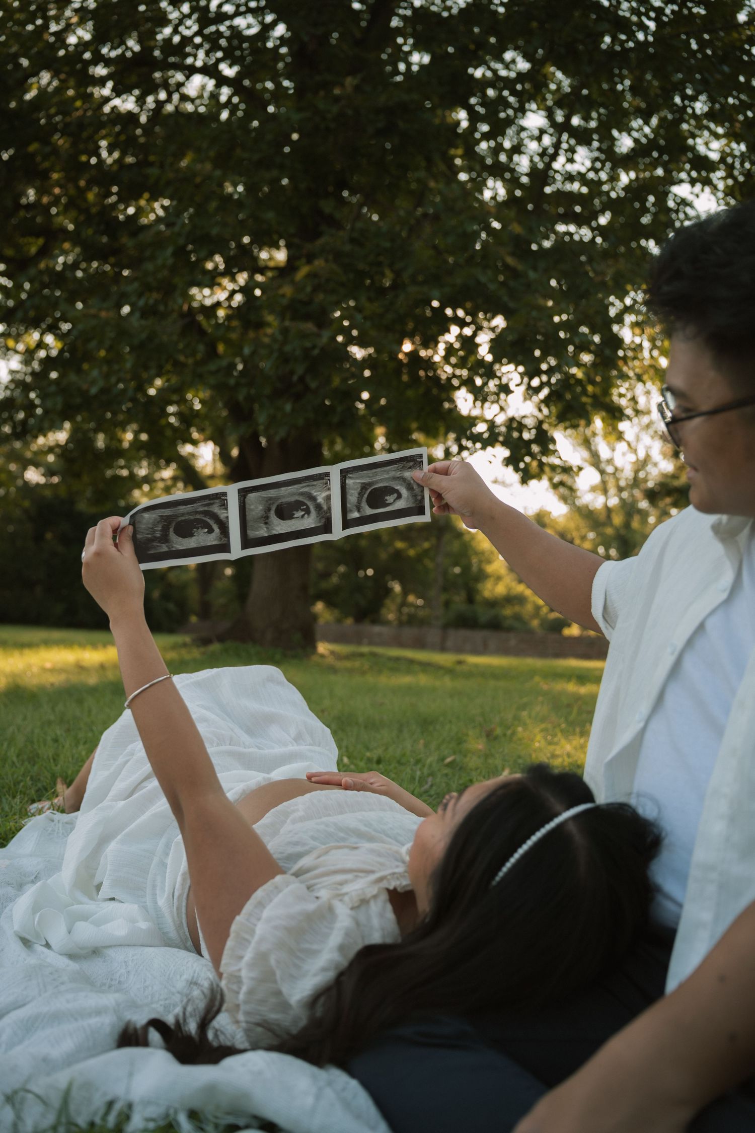 Pregnant couple lying on grass in park holding ultrasound photos, woman in white dress with partner wearing glasses.