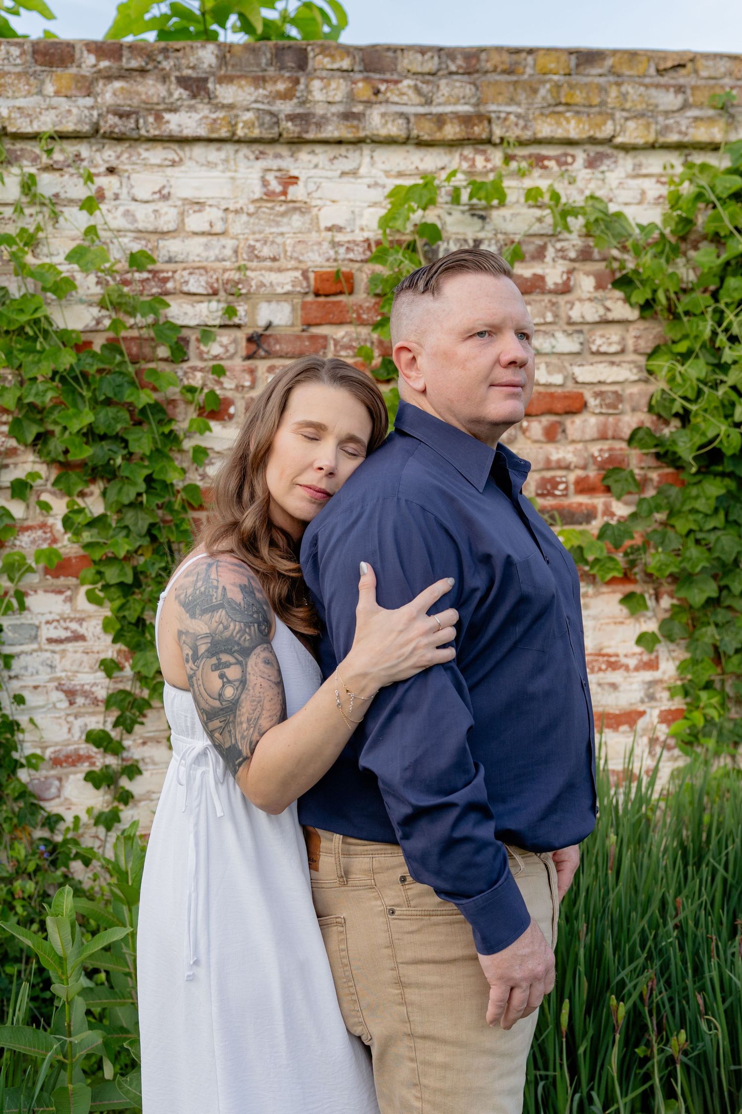Couple posing together in front of an ivy-covered brick wall, woman with tattoos in white dress. Taken by Row And Co Photography in FXBG VA
