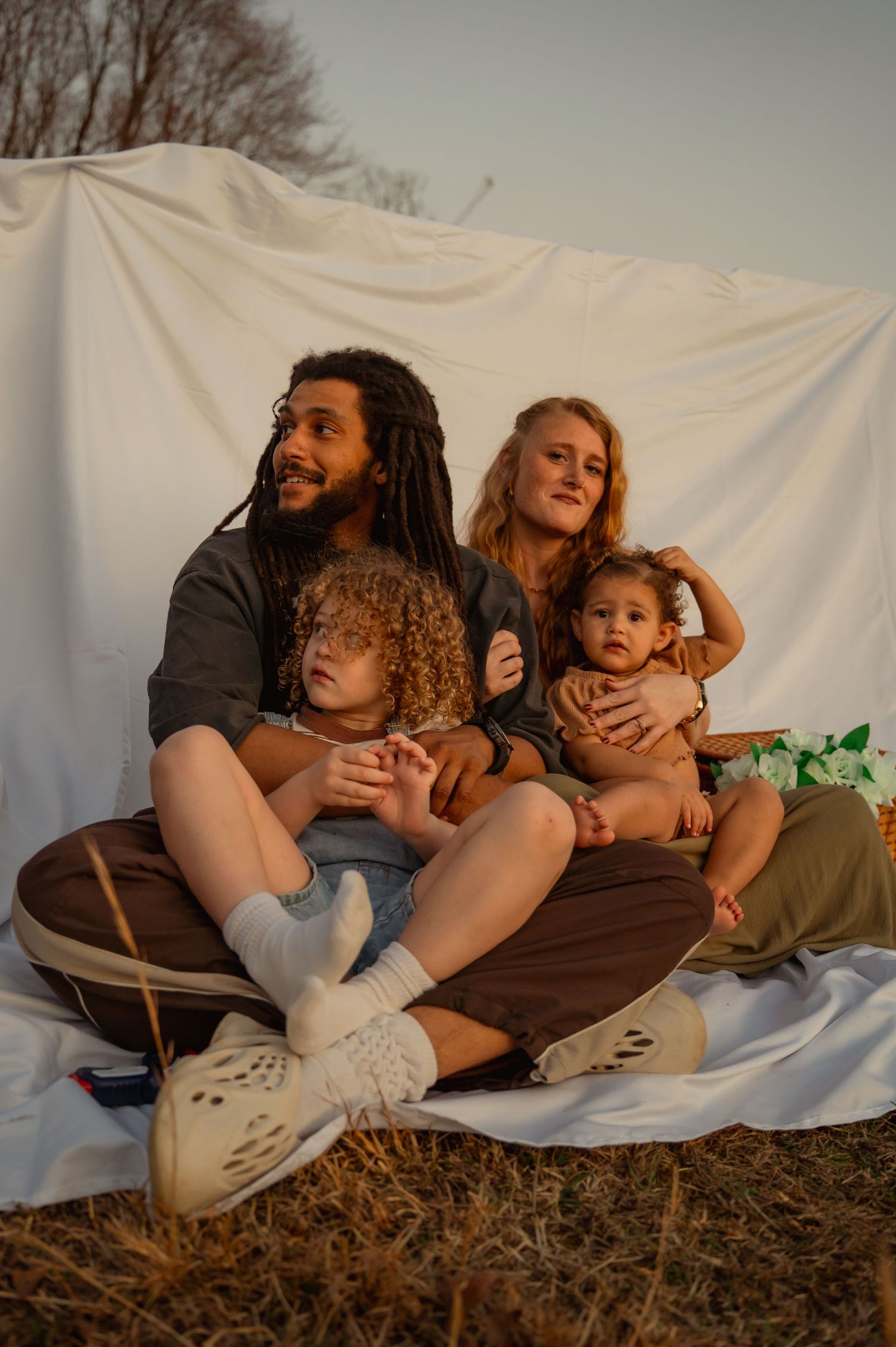 A diverse family of four sits together on a white blanket outdoors, with two young children on their laps at golden hour. Taken by Row&CO