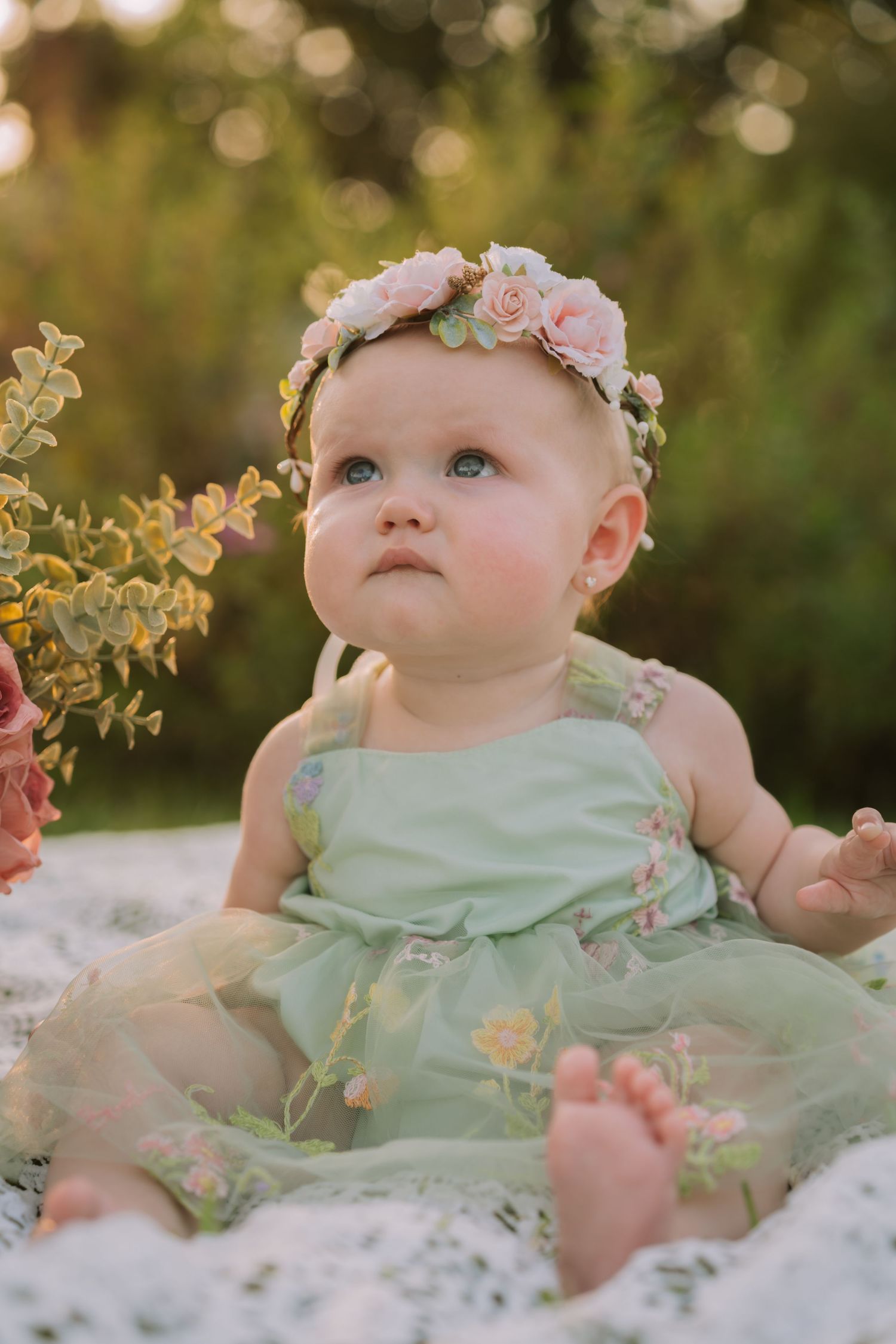 Baby girl in green floral dress and pink rose headband sits outdoors in golden hour light.