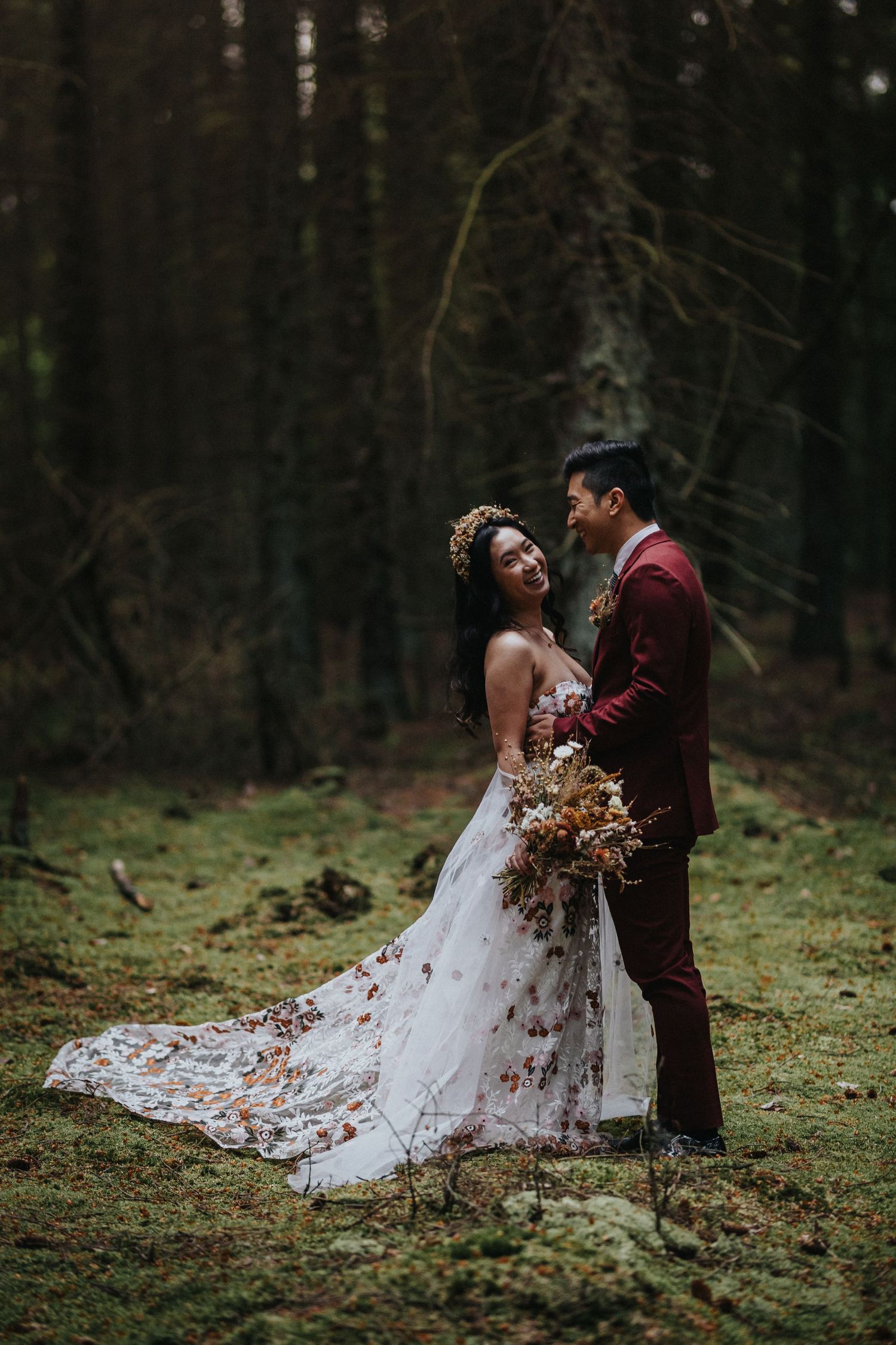 Romantic forest wedding portrait featuring couple in burgundy and white with lace train and autumn bouquet in mossy woods.