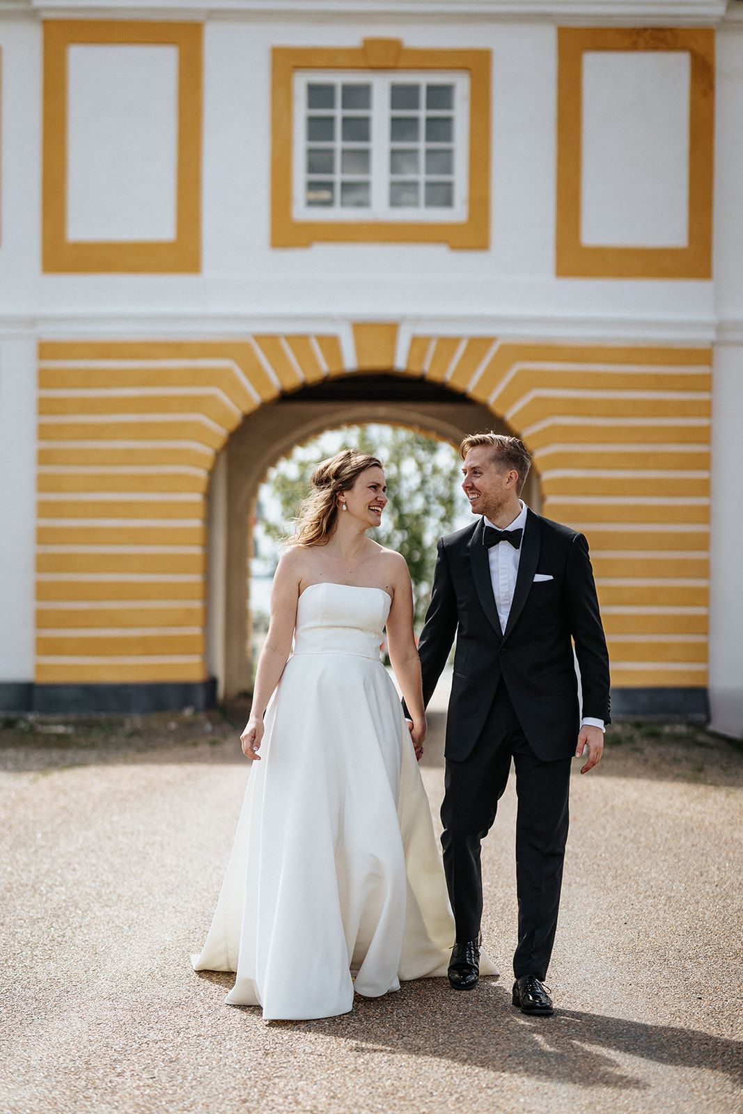 A couple in wedding attire walks together under a yellow arched entrance of a historic building.