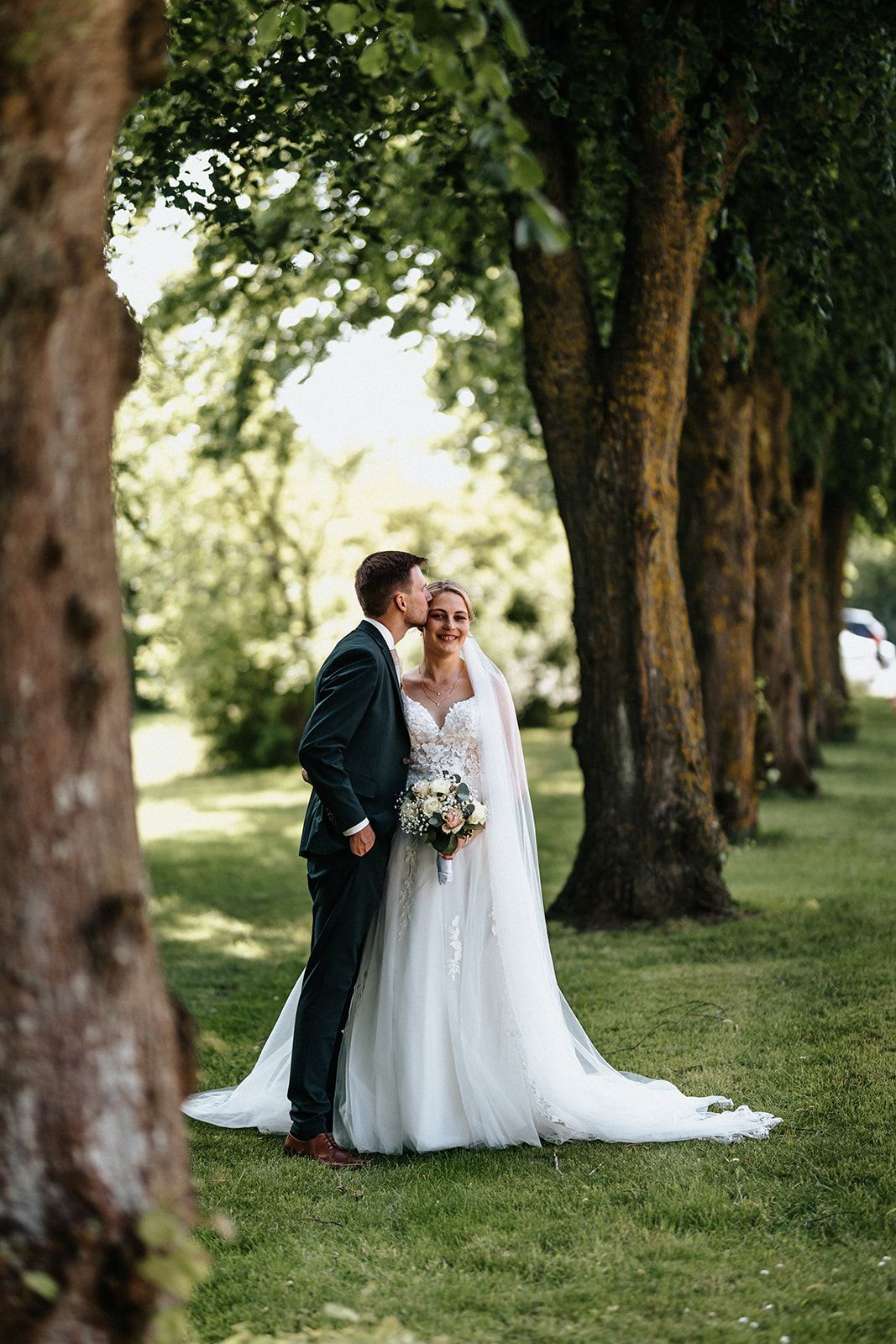 A couple shares a romantic moment under tall trees in a garden setting during their wedding celebration.