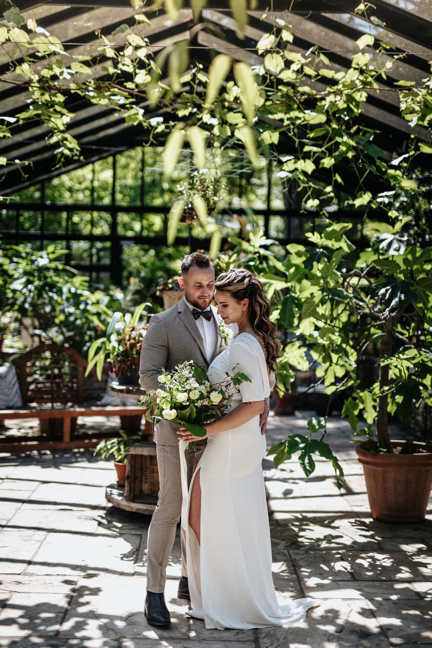 A couple in wedding attire embrace in a sunlit greenhouse with climbing vines and terracotta pots creating a natural backdrop.