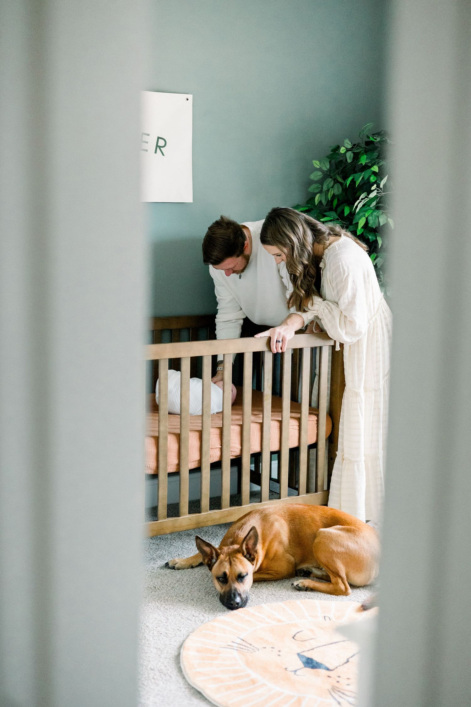 Two people lean over a wooden crib while a brown dog rests on the floor beside a decorative rug. Evans newborn photographer.