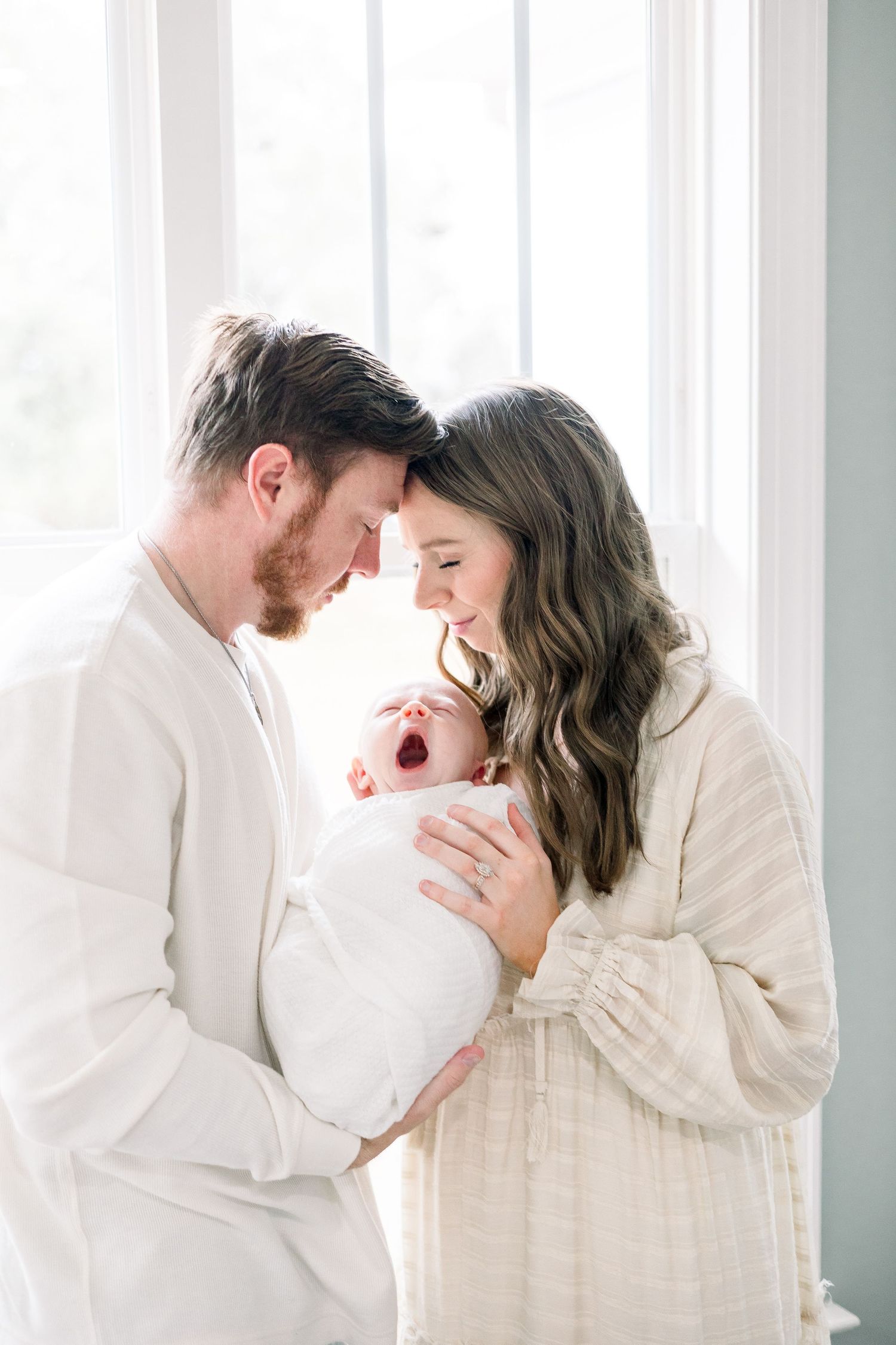 A tender family moment as new parents in cream-colored clothing hold their crying newborn baby by a bright window. Evans newborn photographe