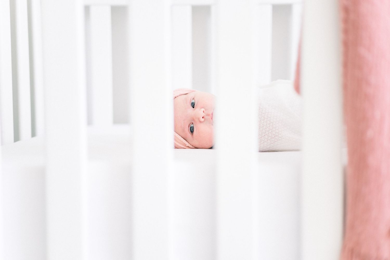 A baby peeks through white crib slats in a soft pink nursery setting. best newborn photographer near me