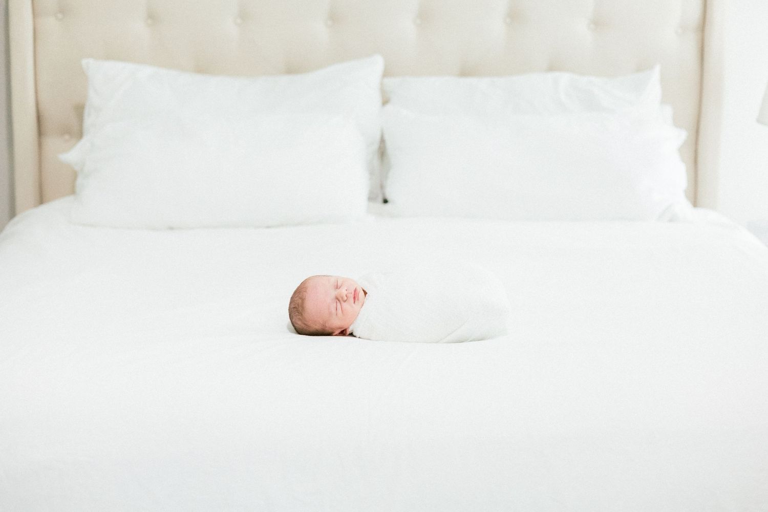 A sleeping newborn lies centered on a pristine white bed with soft white pillows at the headboard. Evans newborn photographer.