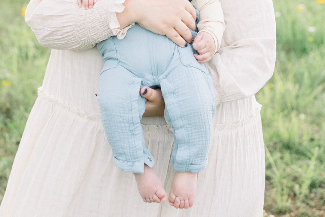 A person in a white dress sits on grass holding a baby wearing light blue pants and a cream colored top. Augusta family photographer