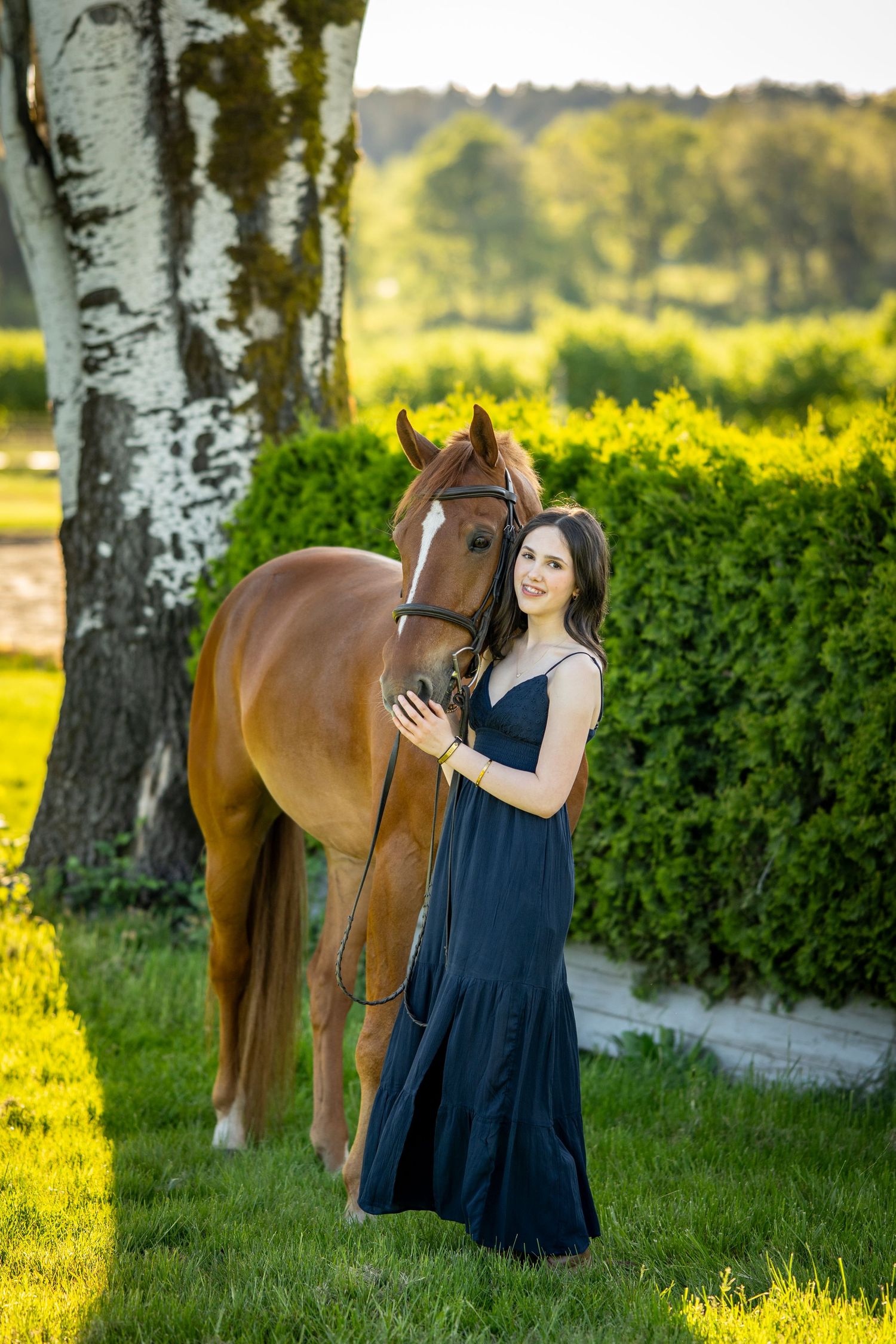 A young woman in a navy blue dress stands beside a chestnut horse in a lush green garden on a sunny day.