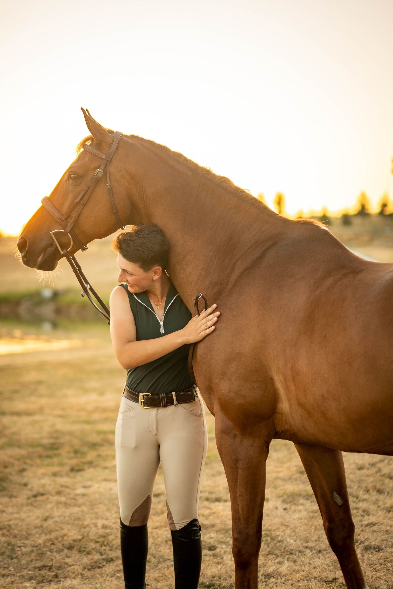A woman in equestrian attire embraces a chestnut horse at golden hour in an open field.