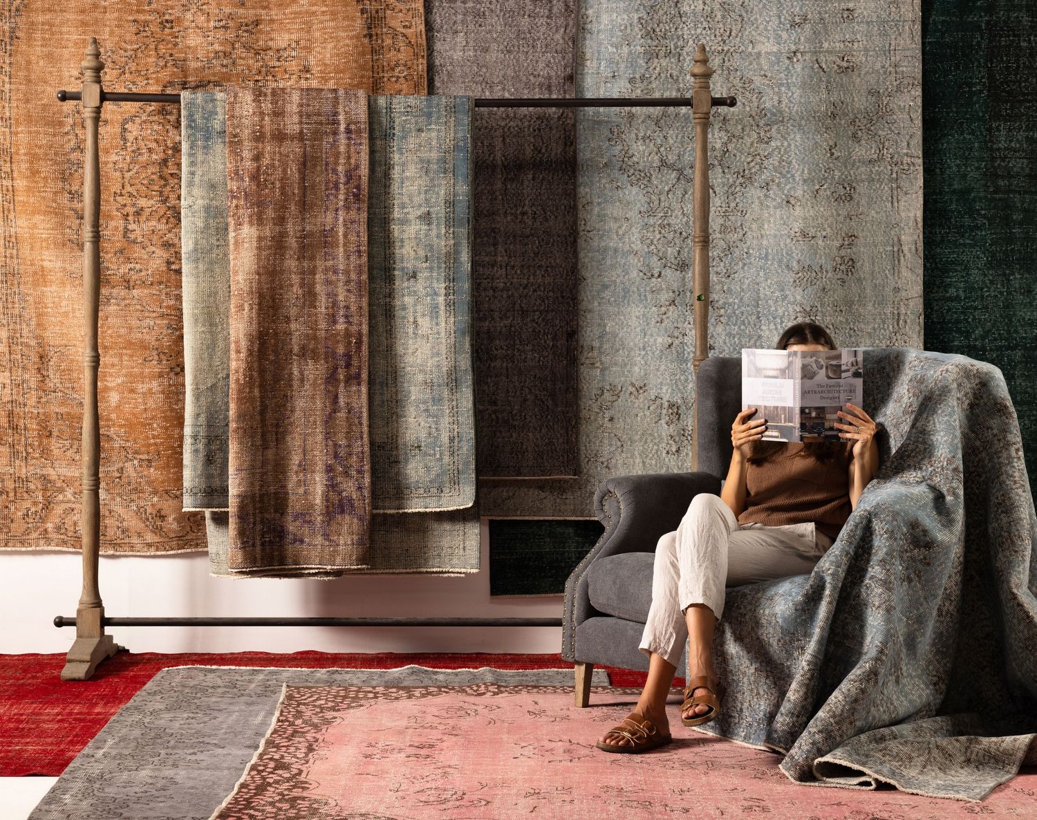 A cozy reading nook with wooden cross wall decor, woven rugs, and someone relaxing in a grey armchair with a blanket.