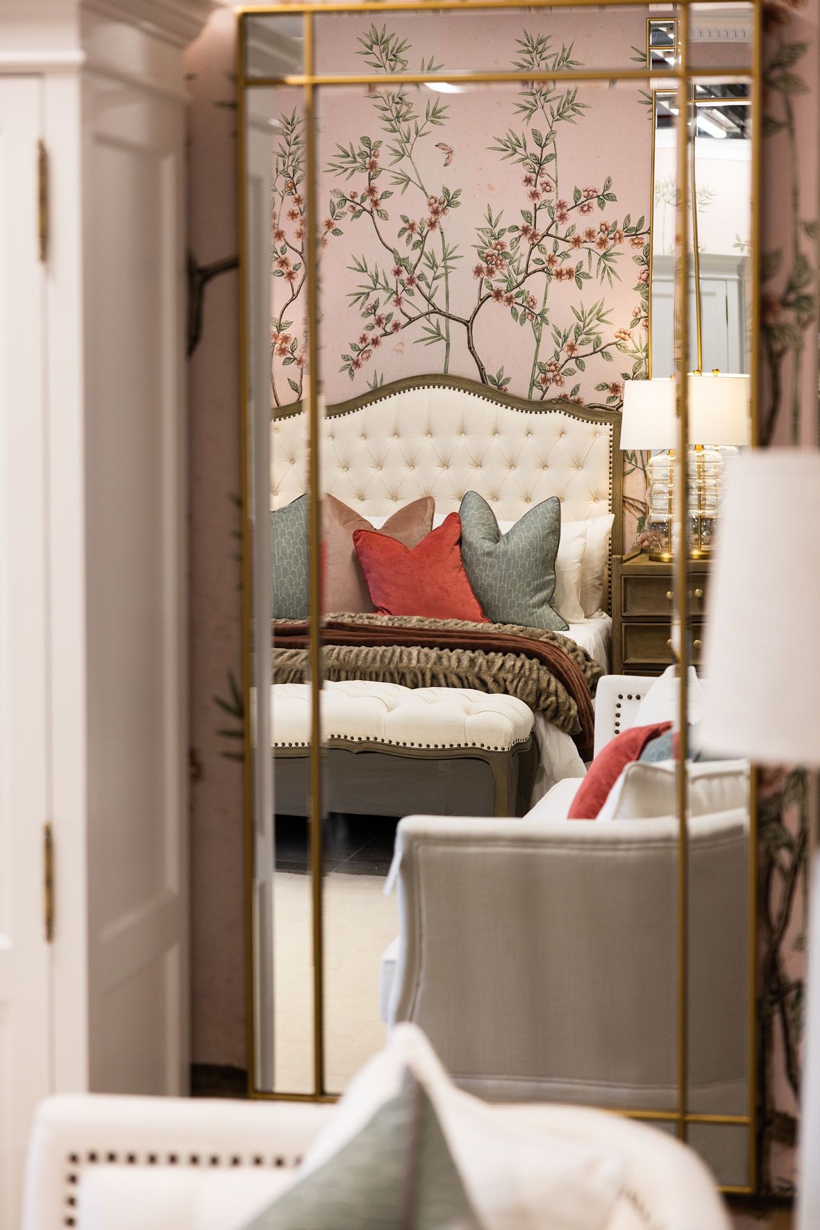 Elegant bedroom glimpse through mirror frame showing floral wallpaper and tufted headboard with decorative pillows.