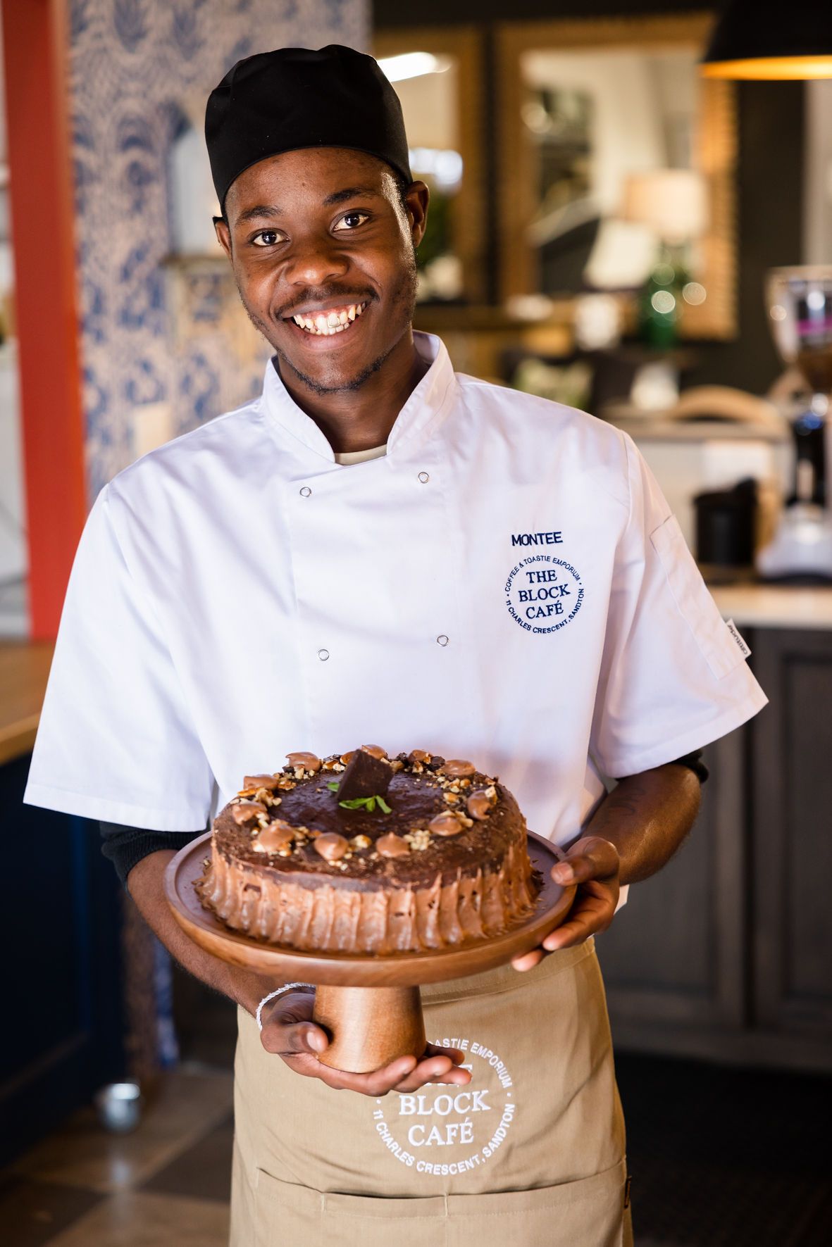 Chef in white uniform and hat proudly displays a large chocolate dessert in a restaurant kitchen.