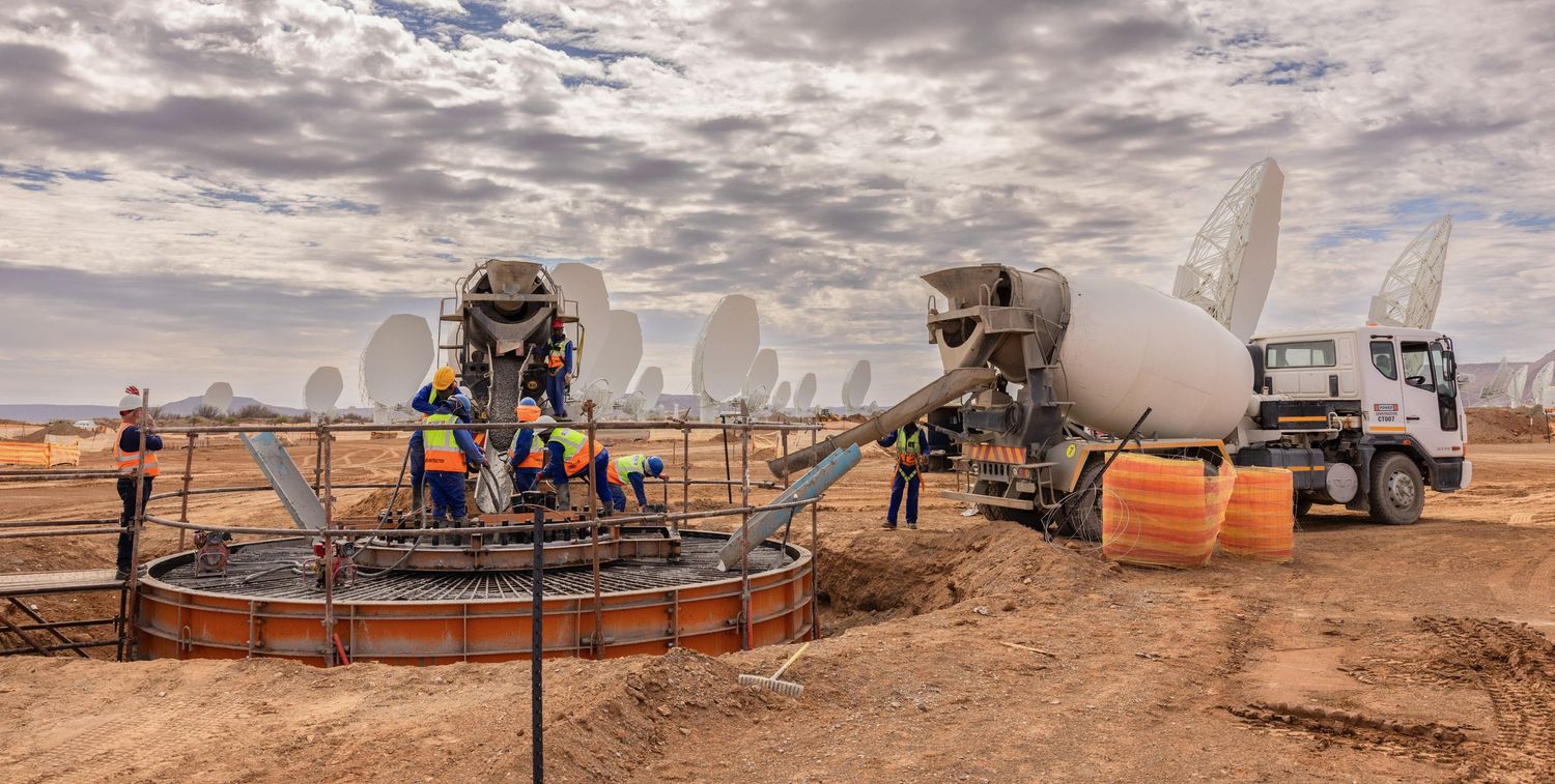 Construction site with cement mixers and workers installing satellite dish equipment under cloudy skies.