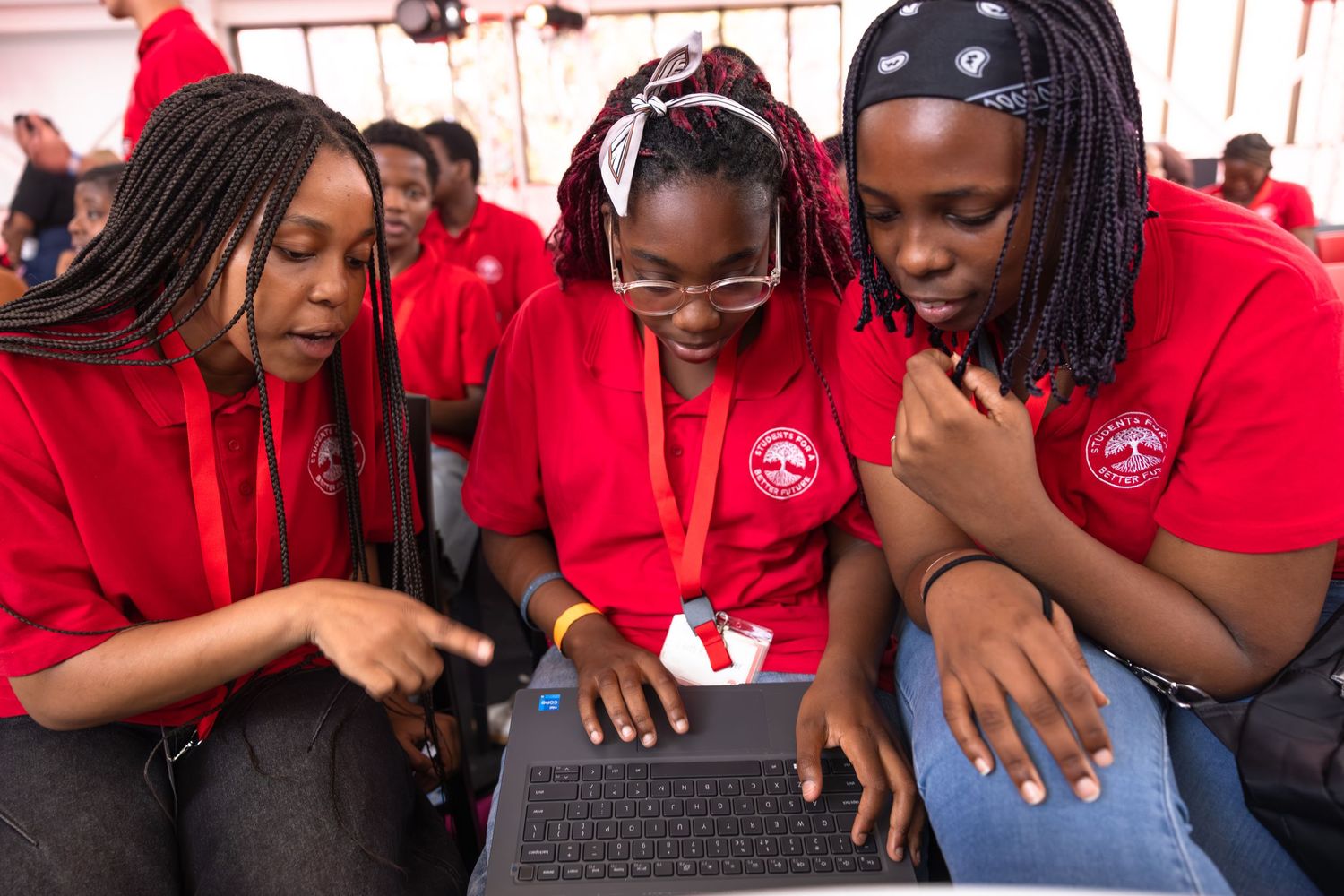 Three students in red shirts collaborate while looking at a laptop computer together.