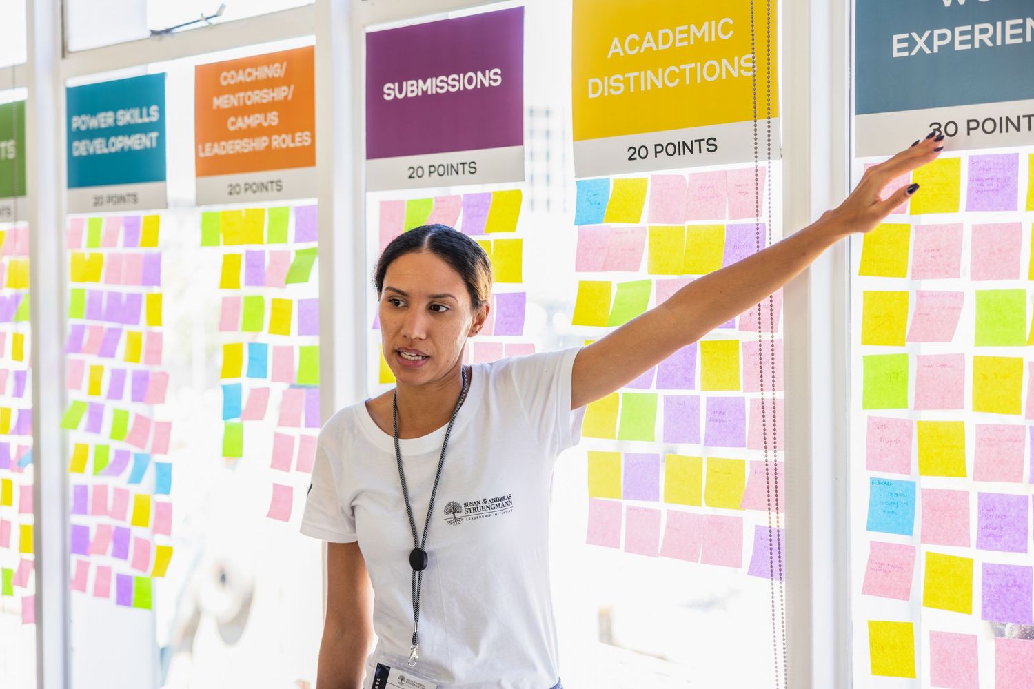 Student points to colorful sticky notes arranged on wall columns with academic categories.