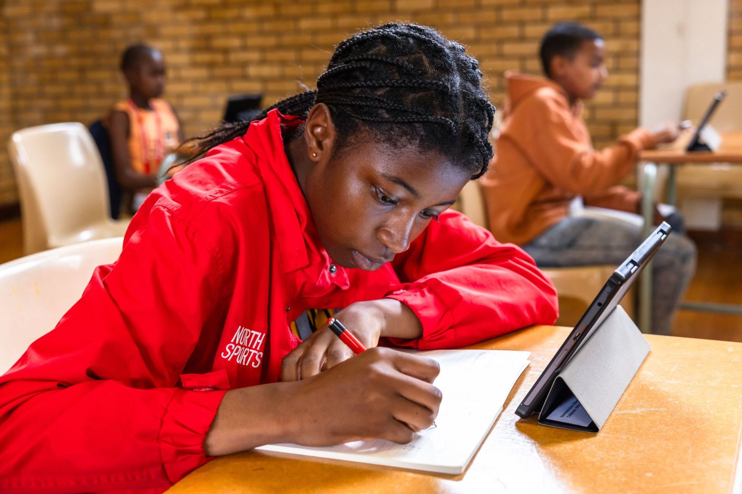 Student in red jacket focuses on writing notes while using tablet device in classroom.